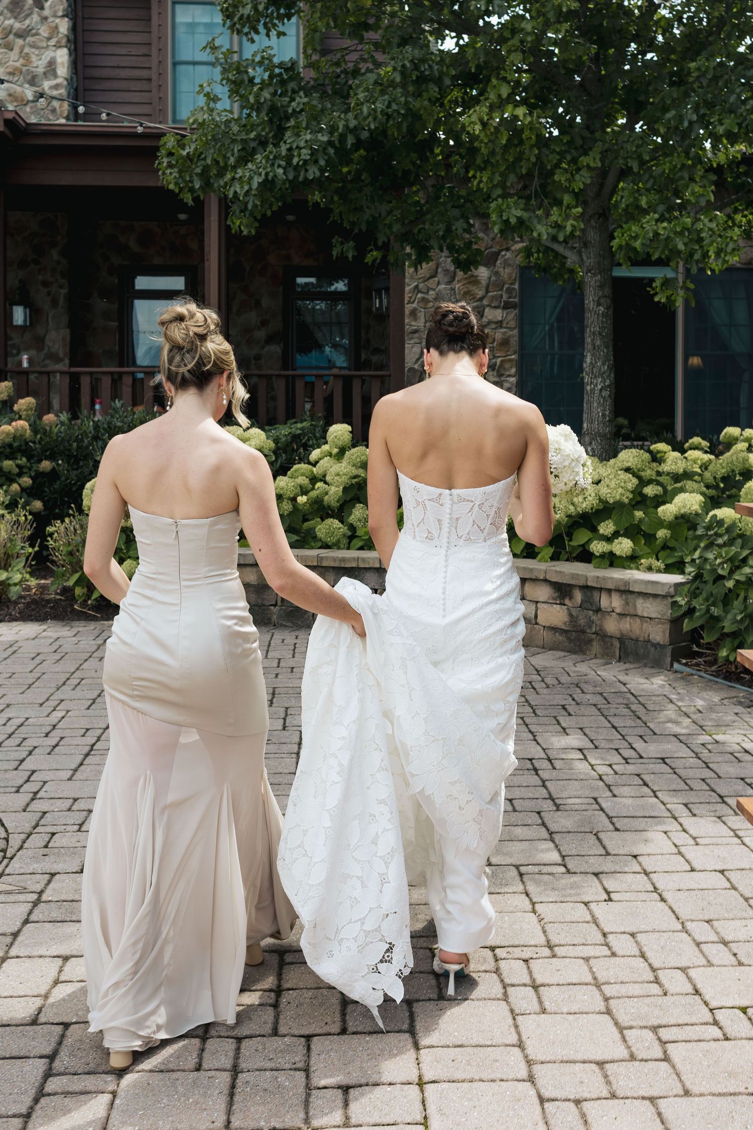 Bride in white lace gown walks with bridesmaid in champagne dress on stone path, both strapless, viewed from behind.