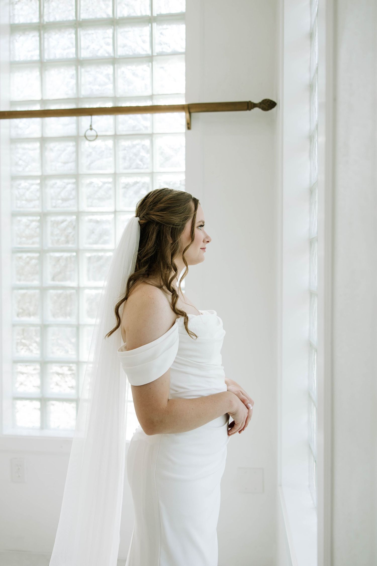 A bride in a white off-shoulder wedding dress stands by bright windows in an elegant indoor setting.