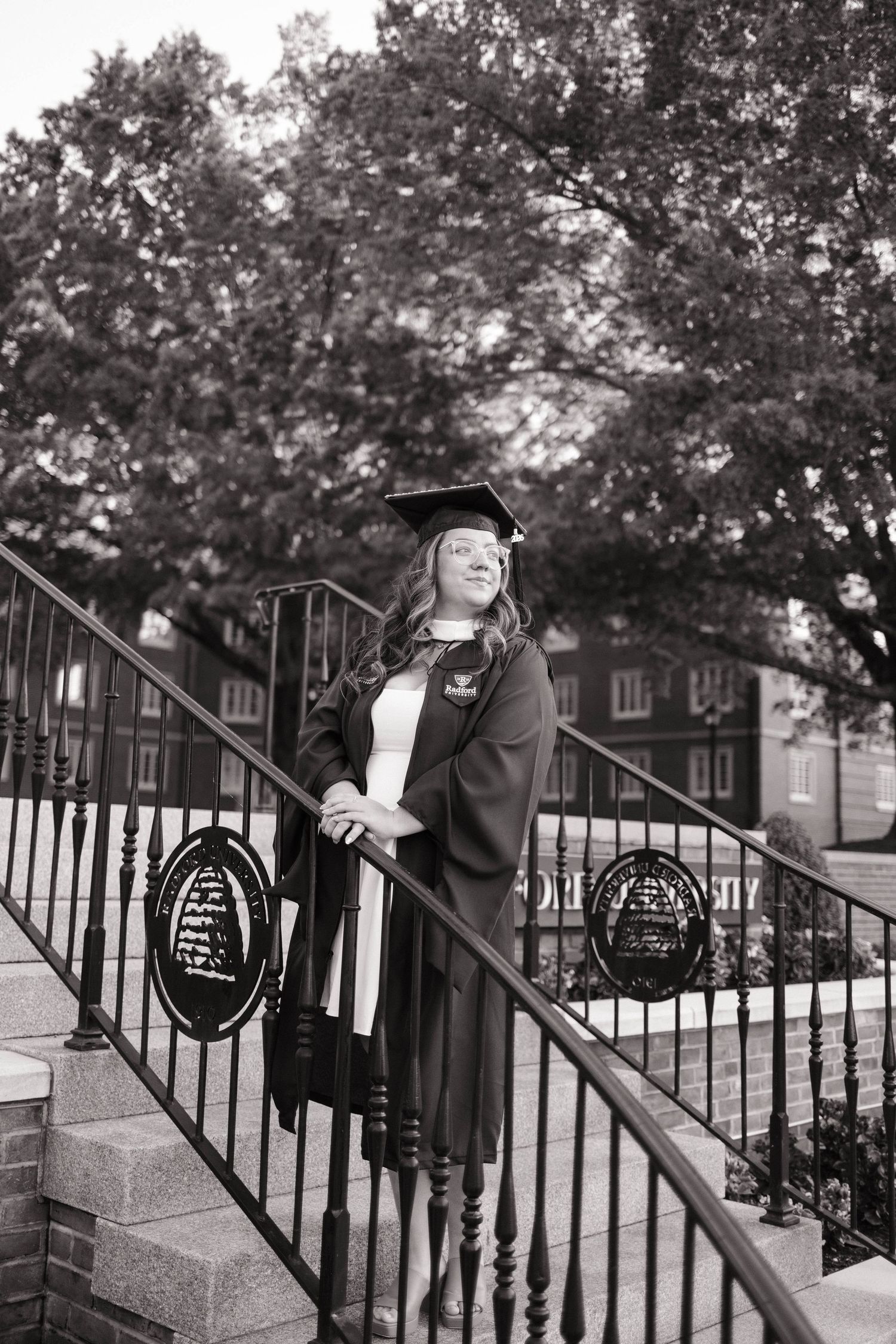 Black and white photo of a young woman in graduation cap and gown posing on outdoor steps with iron railings.