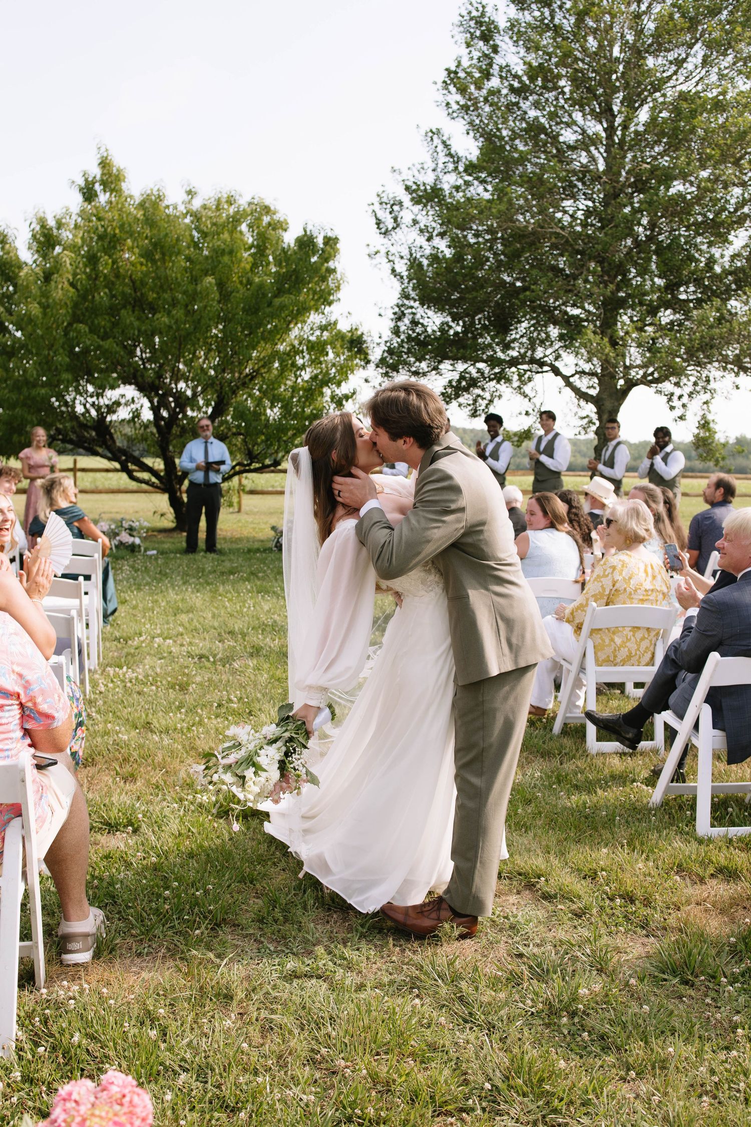 Bride and groom share first kiss at outdoor wedding ceremony on green lawn with seated guests.