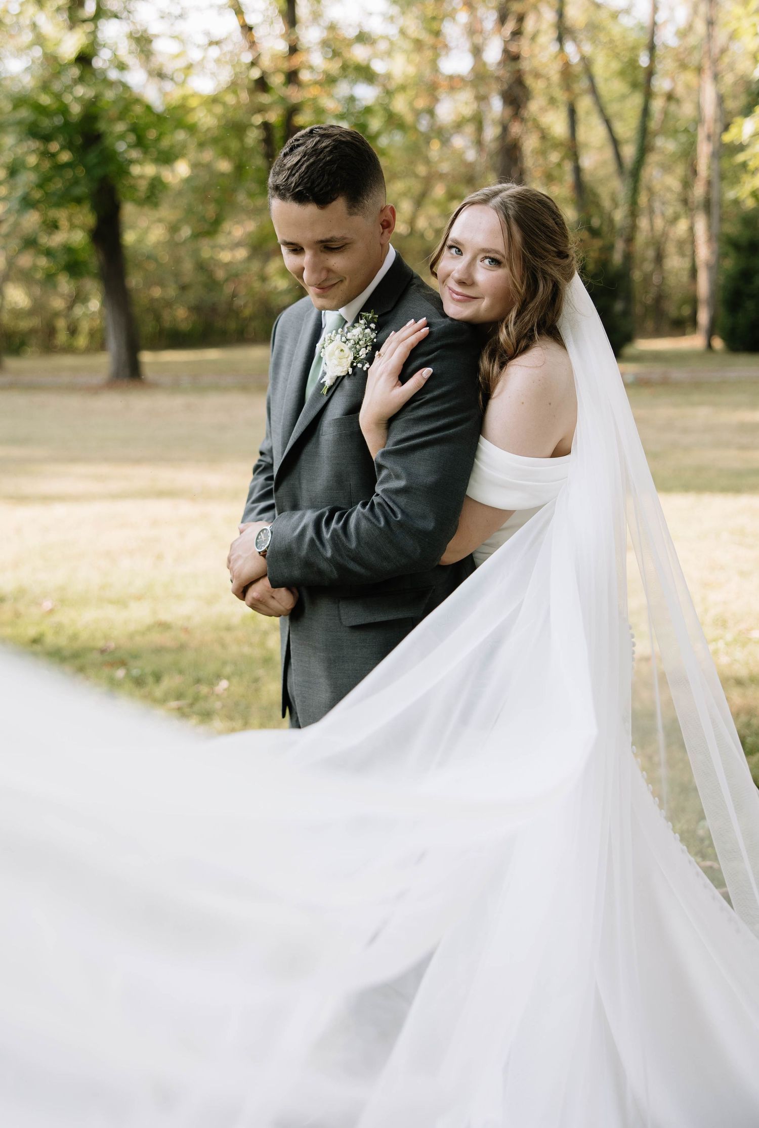 A romantic wedding couple embraces outdoors in an autumn setting as the bride's flowing white veil catches the breeze.