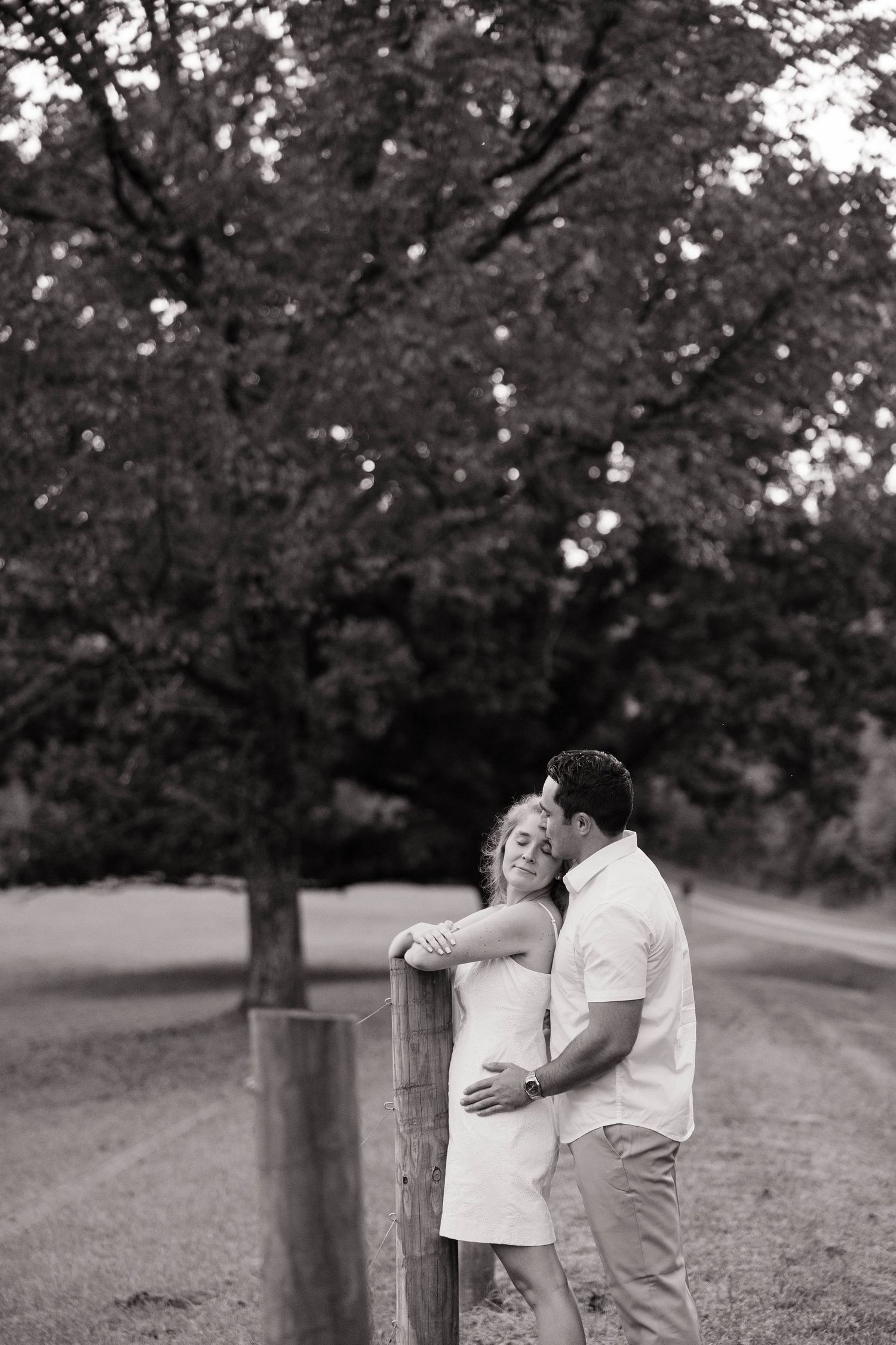 Black and white photo of a couple sharing romantic moments along a tree-lined path.