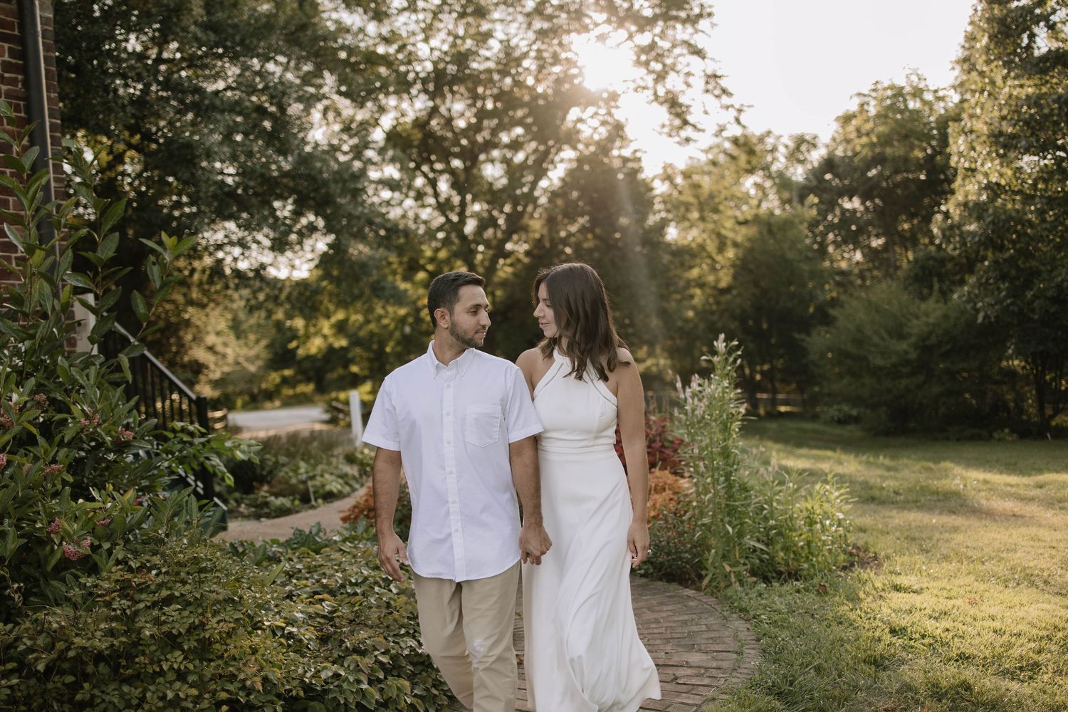 A couple walks hand in hand at sunset with trees and foliage in the background.