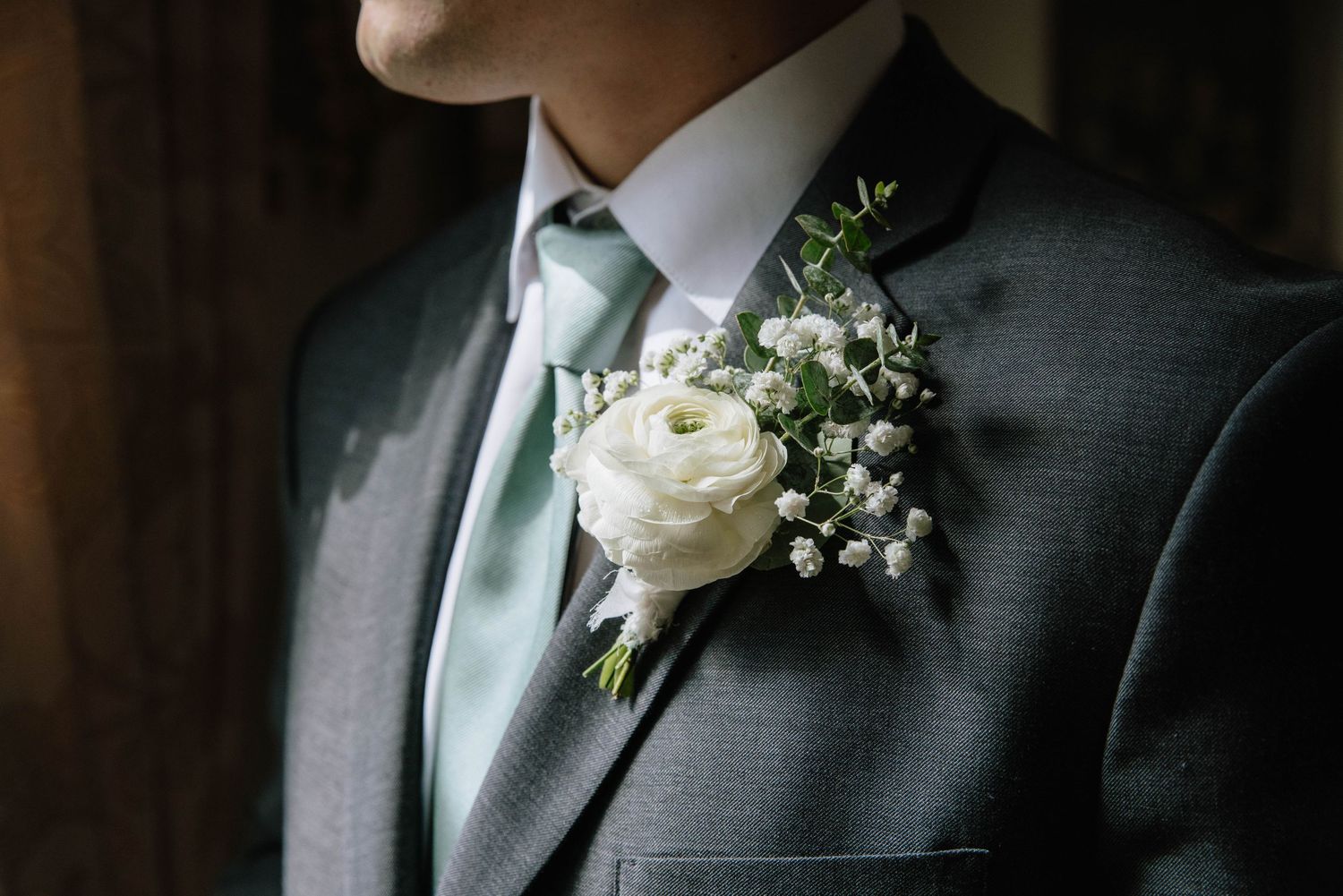 A close-up detail of a formal grey suit with a white boutonniere and green tie at a wedding ceremony.