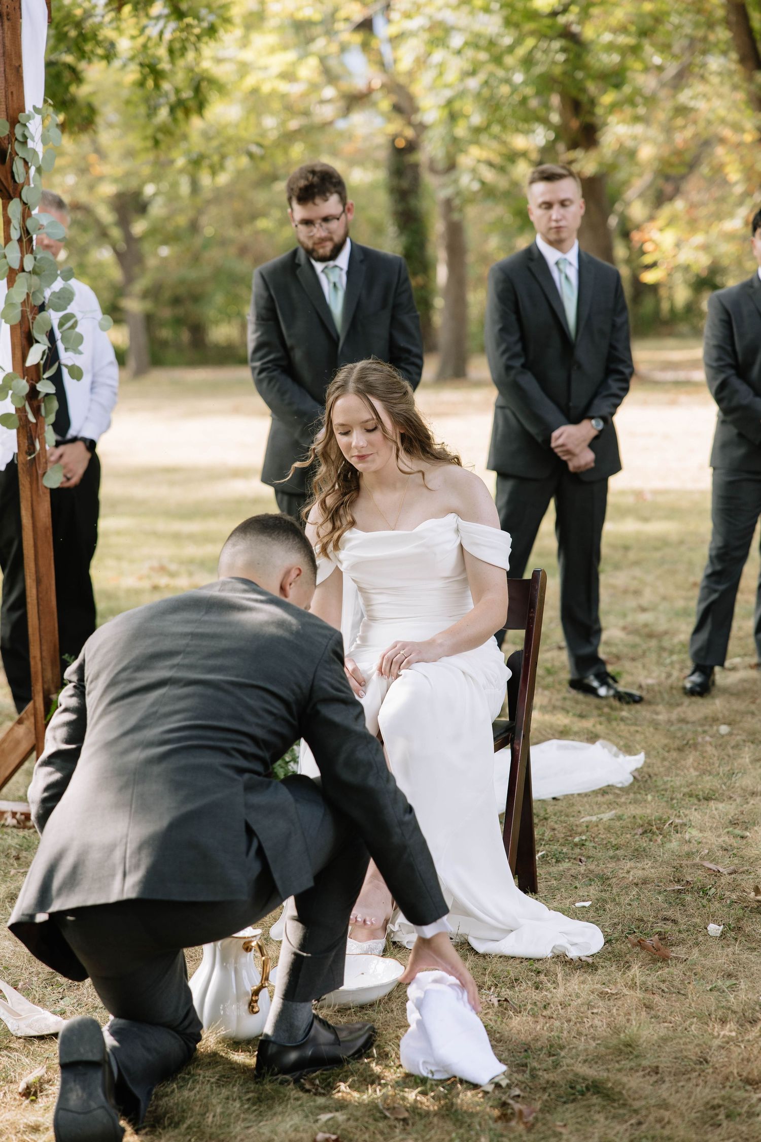 Groomsmen assist with bride's shoe during wedding tradition at outdoor ceremony.