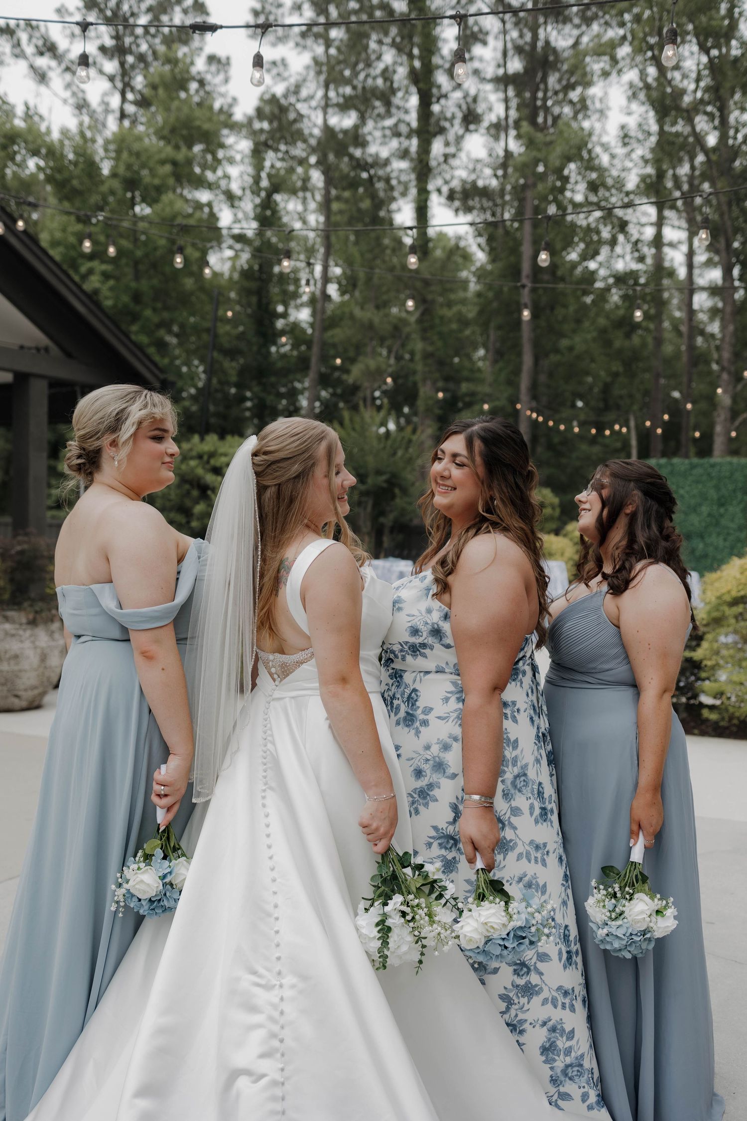 Bride in white dress poses with bridesmaids wearing light blue dresses under string lights.