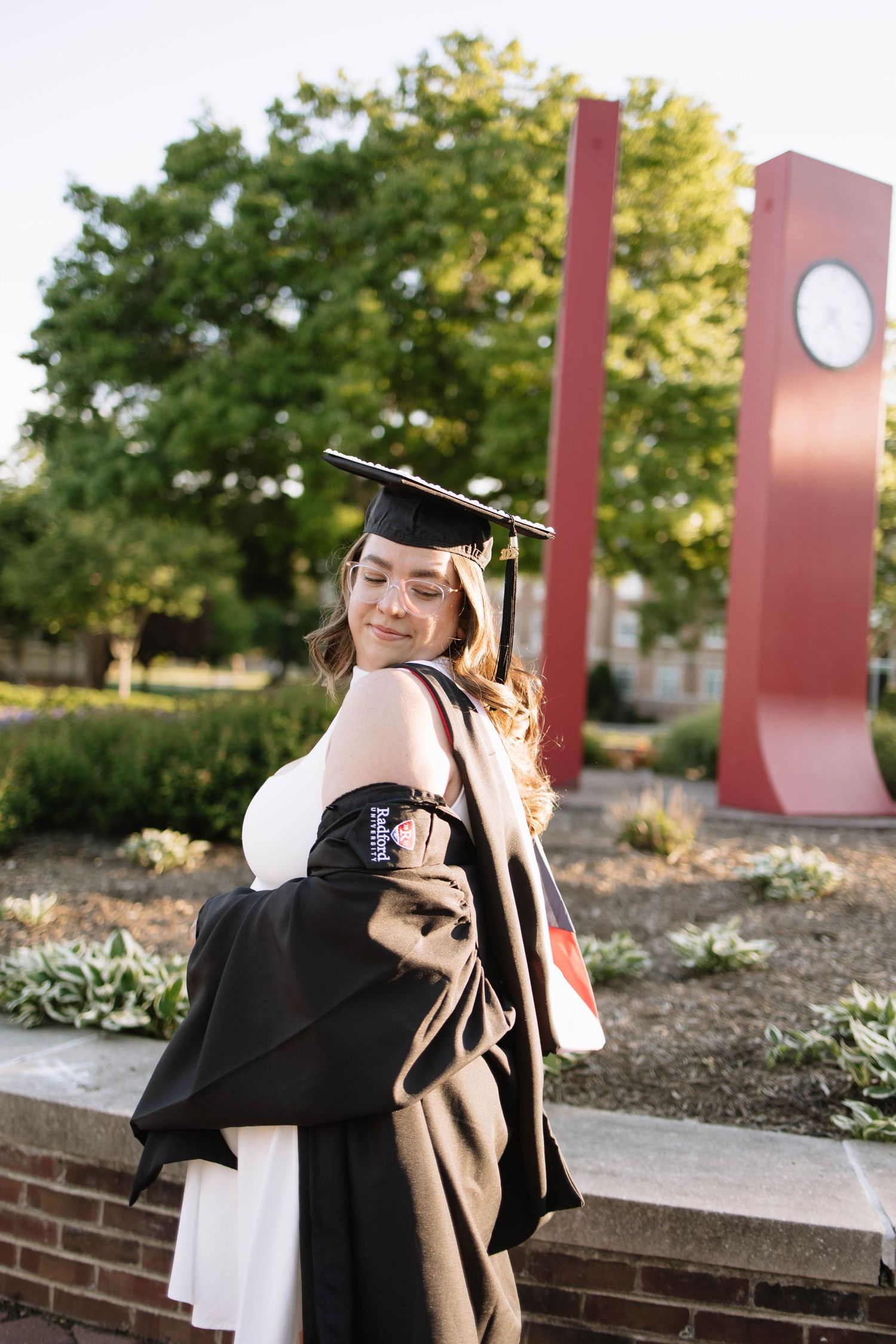 A young woman in a black graduation cap and gown smiles over her shoulder outdoors near a red campus monument.