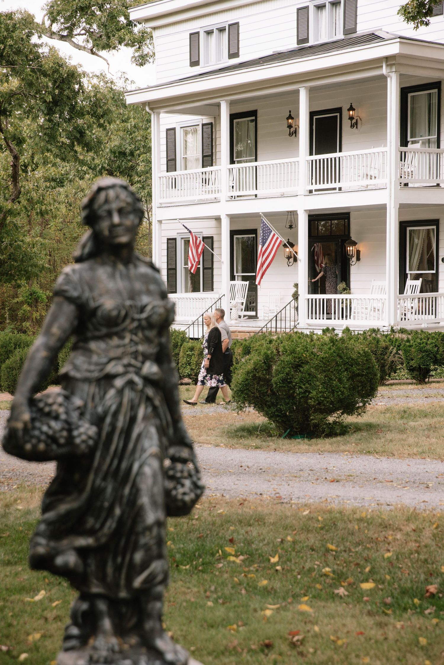 White colonial-style house with wraparound porch and American flags viewed through decorative garden statue silhouettes.