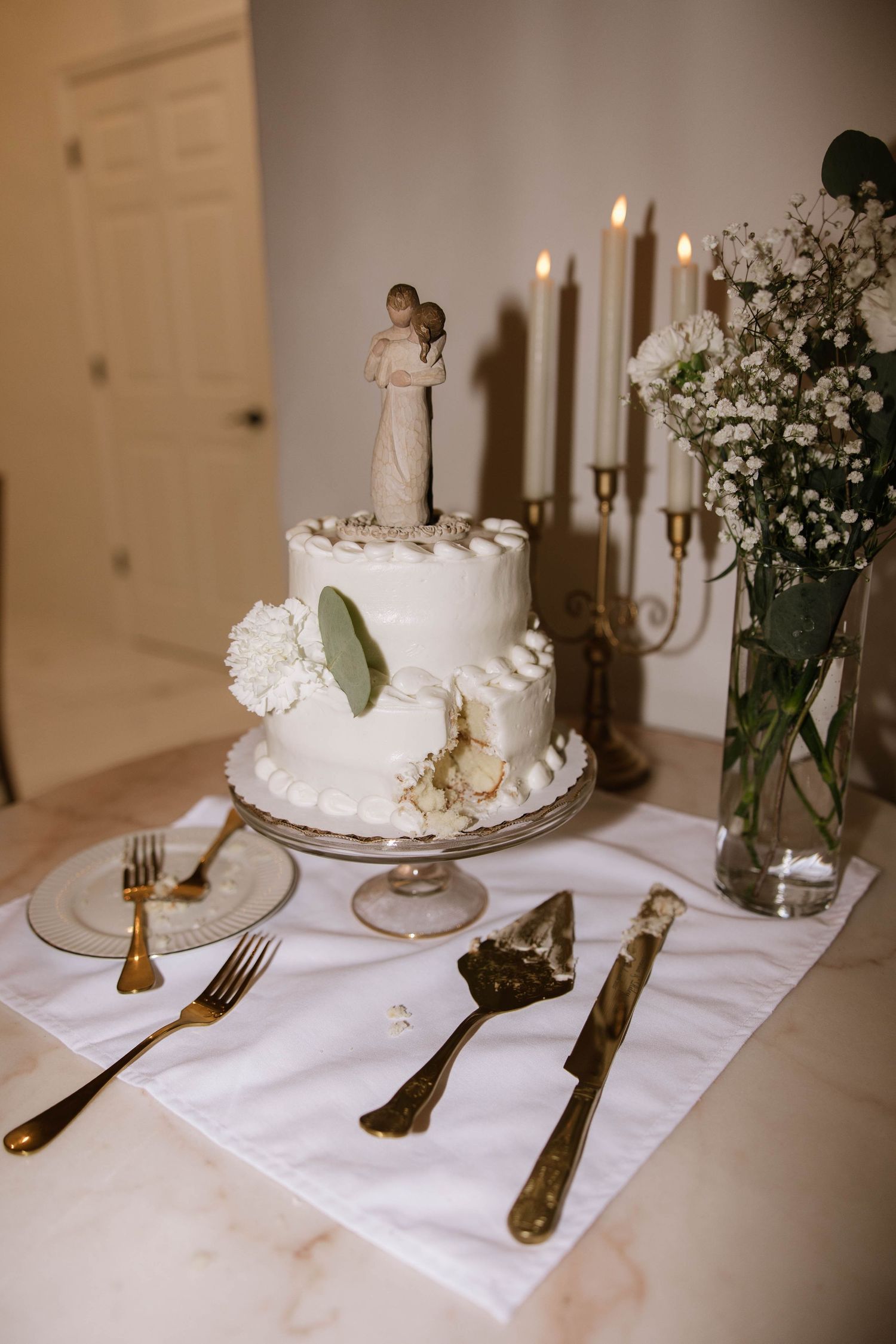 Elegant white wedding cake display with gold utensils and candlesticks on a white tablecloth.