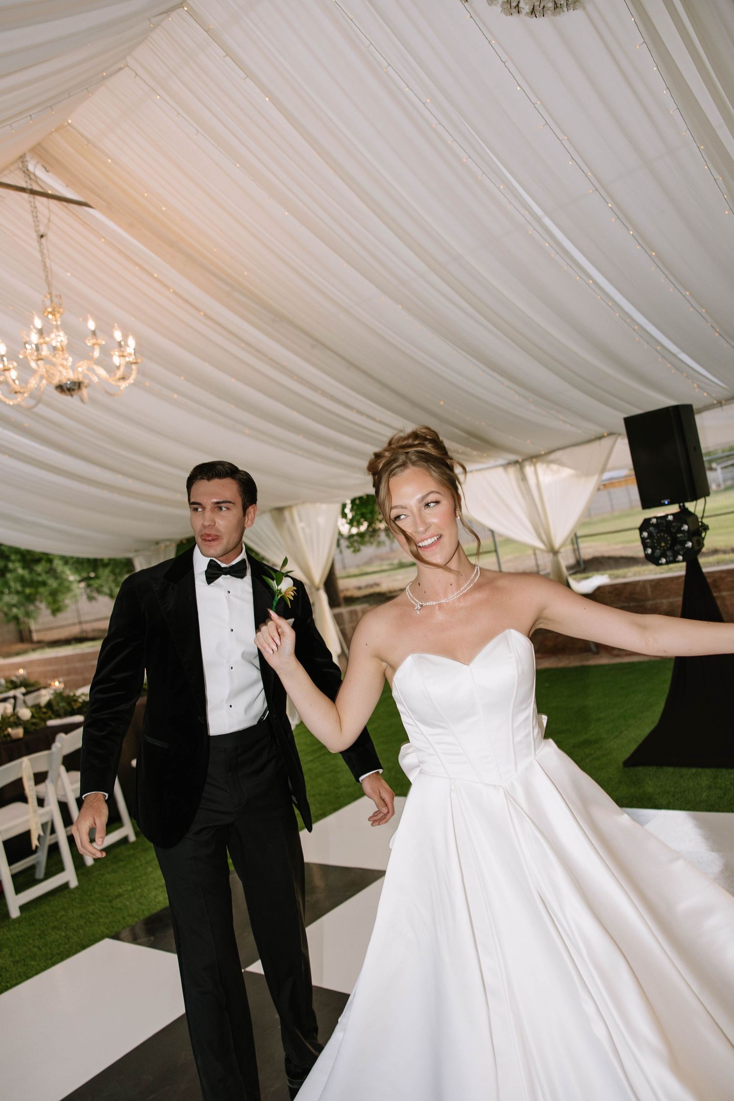 Bride in white strapless gown and groom in black tuxedo entering wedding reception under elegant draped tent.