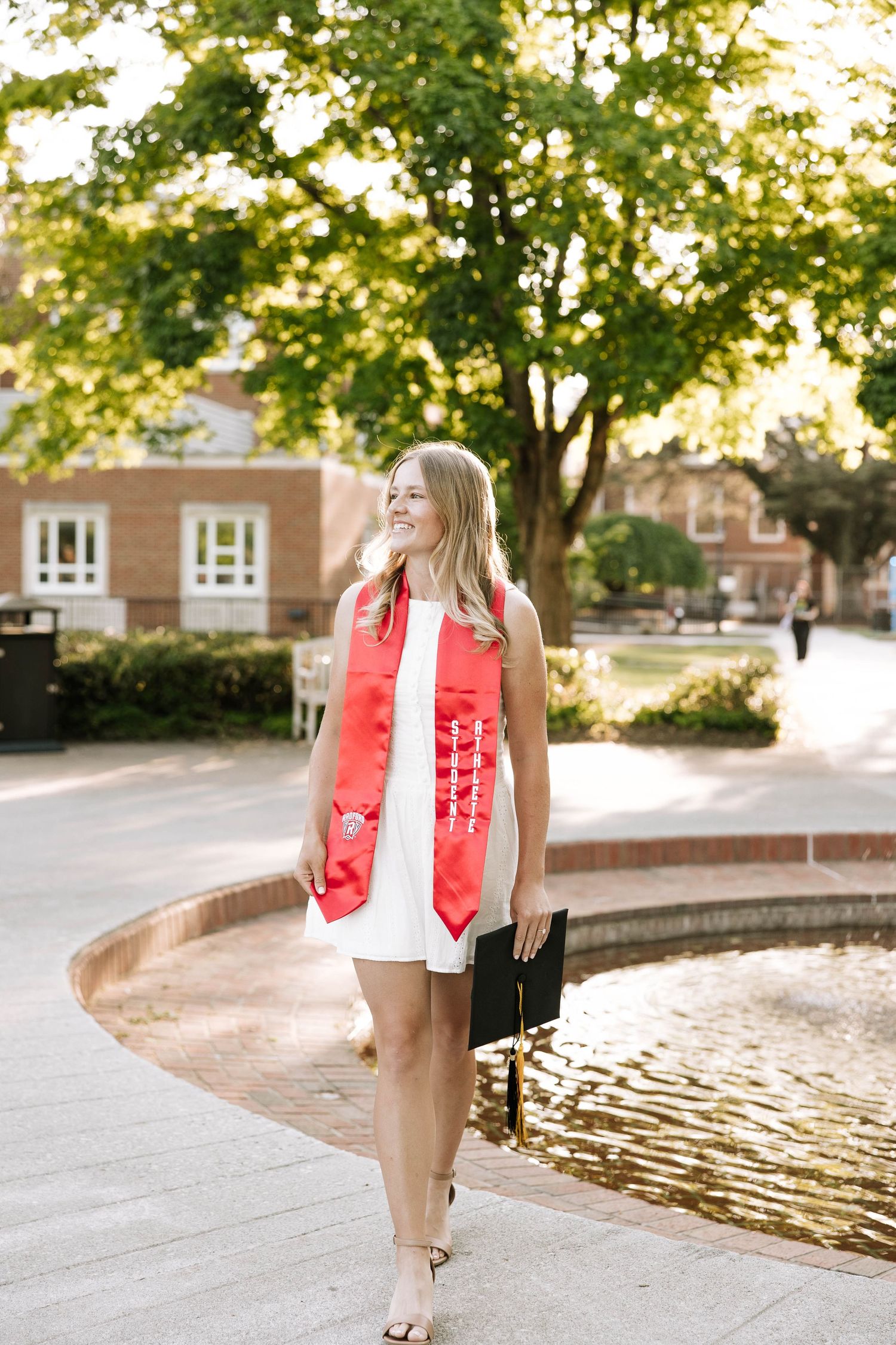 Smiling graduate in white dress and red sash holds cap by fountain on sunny campus.