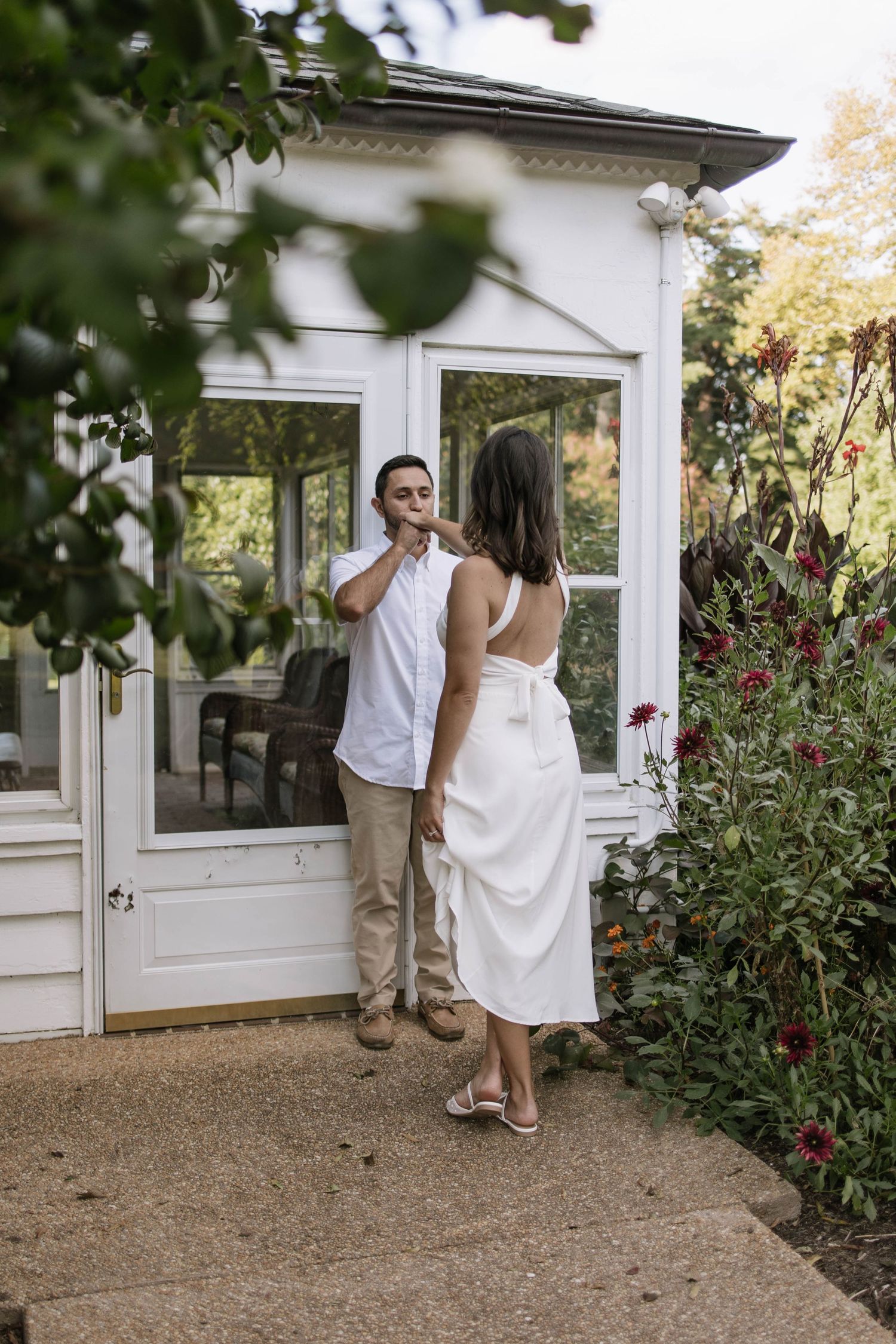 A couple in white clothing shares intimate moments near a white greenhouse with flowering plants.