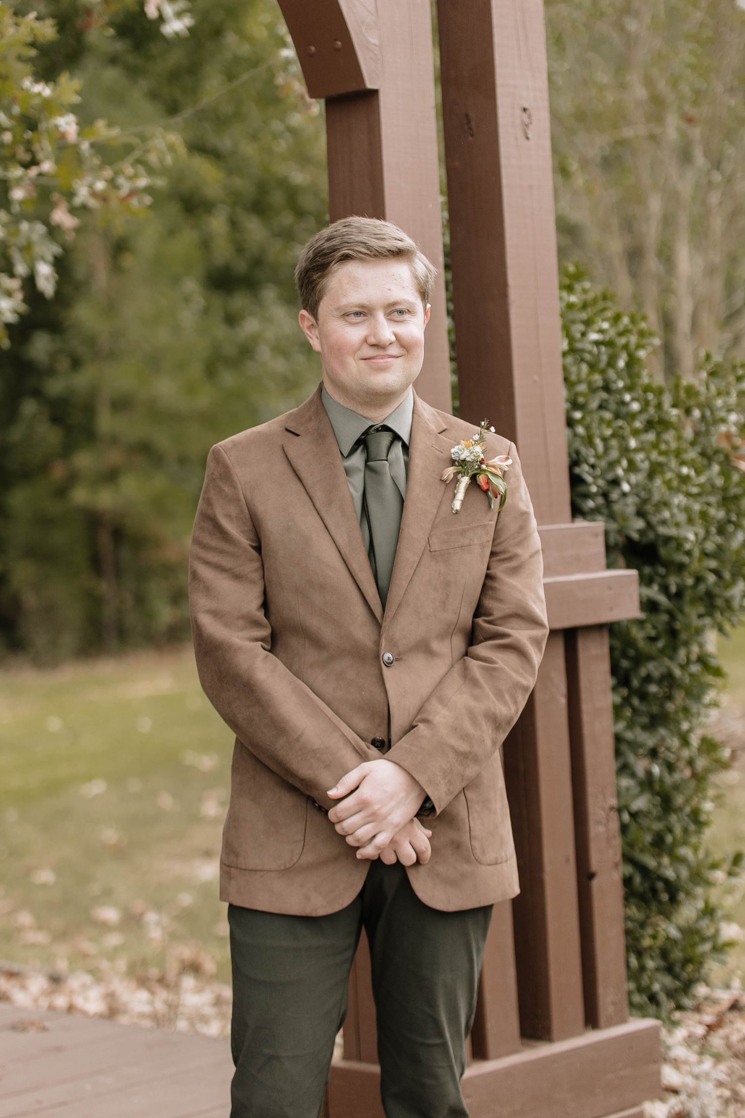 Wedding party member in brown suit and dark tie standing by wooden pergola.