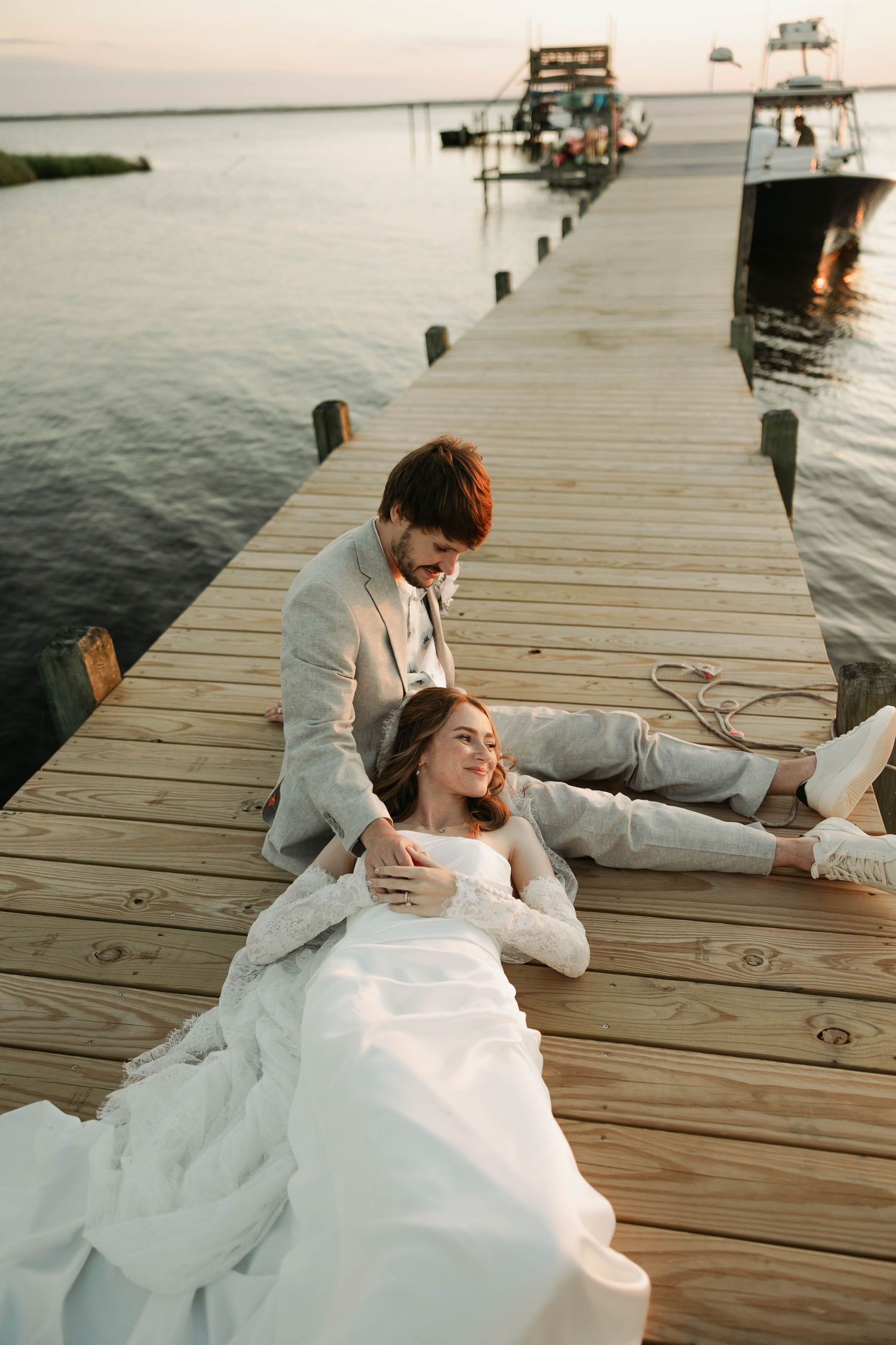 Bride in white gown reclines on wooden dock as groom in gray suit sits beside her at waterfront sunset.