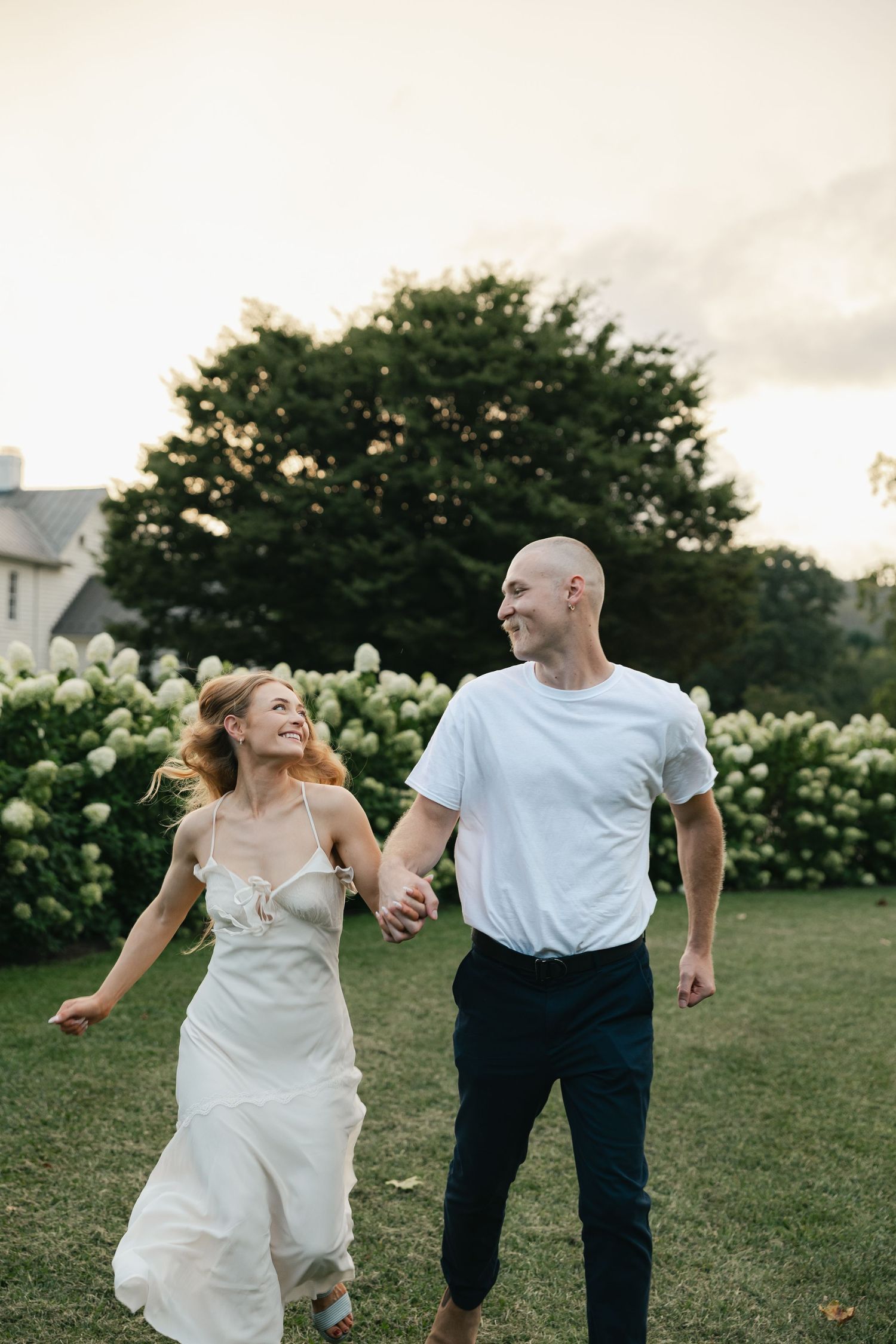 A joyful couple holds hands and runs through a lush green lawn with white hydrangeas at golden hour.