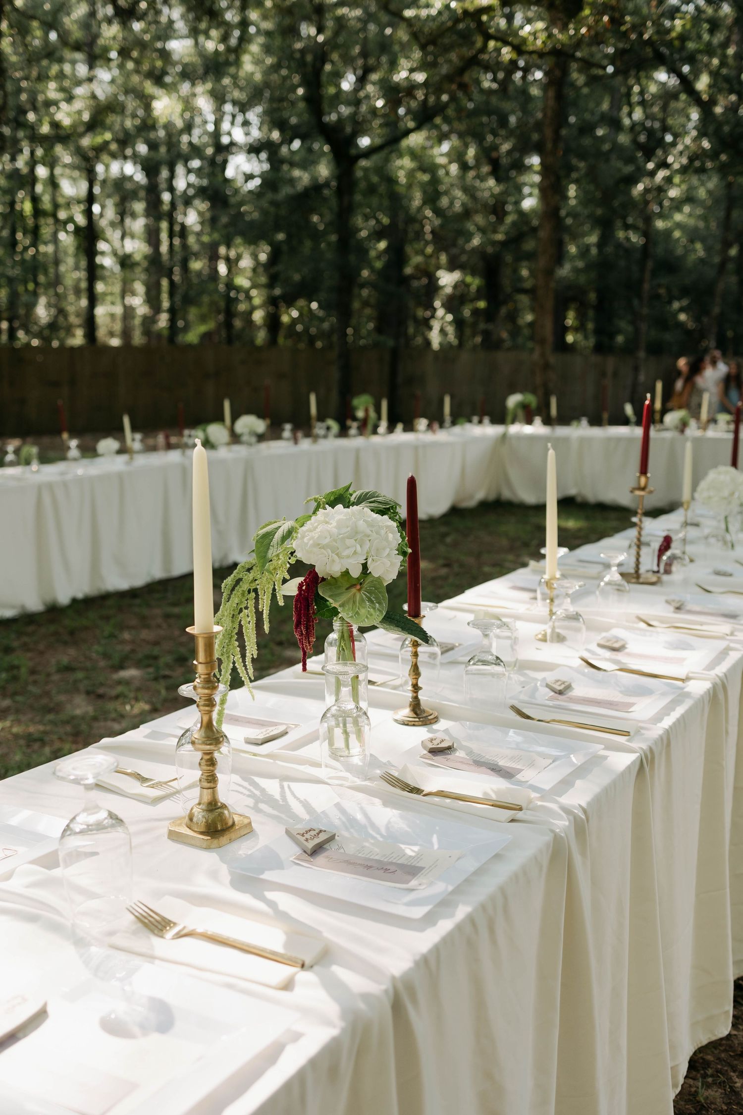 Elegant outdoor wedding reception table with white linens, gold candelabras, and white floral centerpieces in a forest setting.