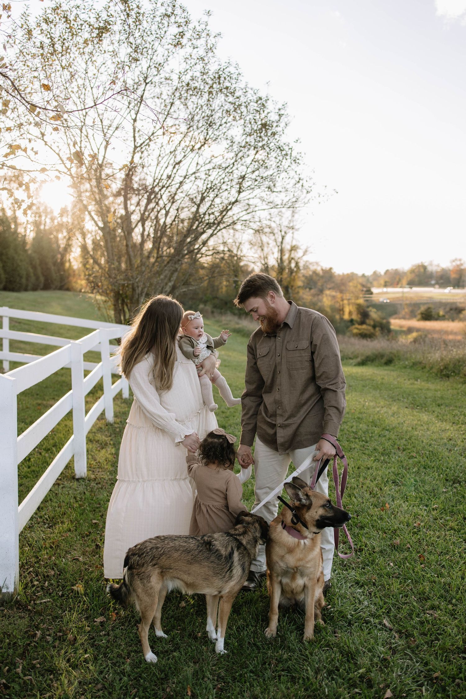 Couple in casual attire standing by a white fence with their dogs at sunset.