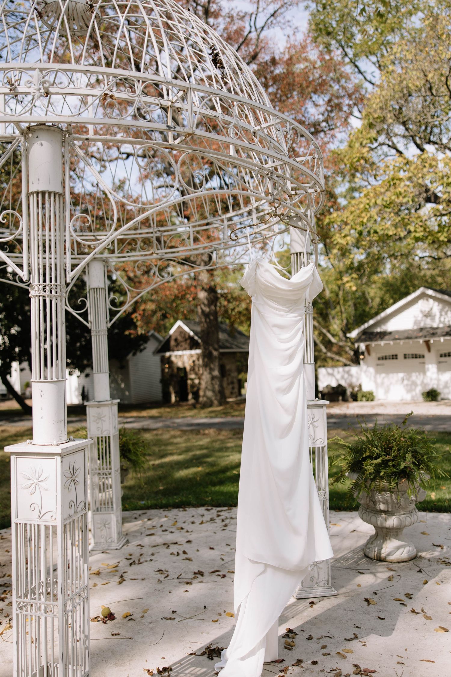 A white wedding dress hangs elegantly from an ornate white metal gazebo surrounded by autumn leaves on the ground.