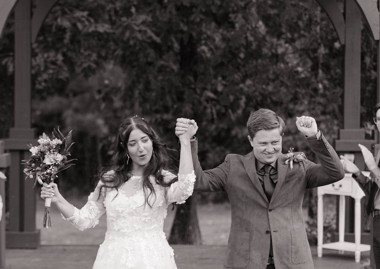 Wedding couple celebrates with joyful gestures under outdoor pavilion.