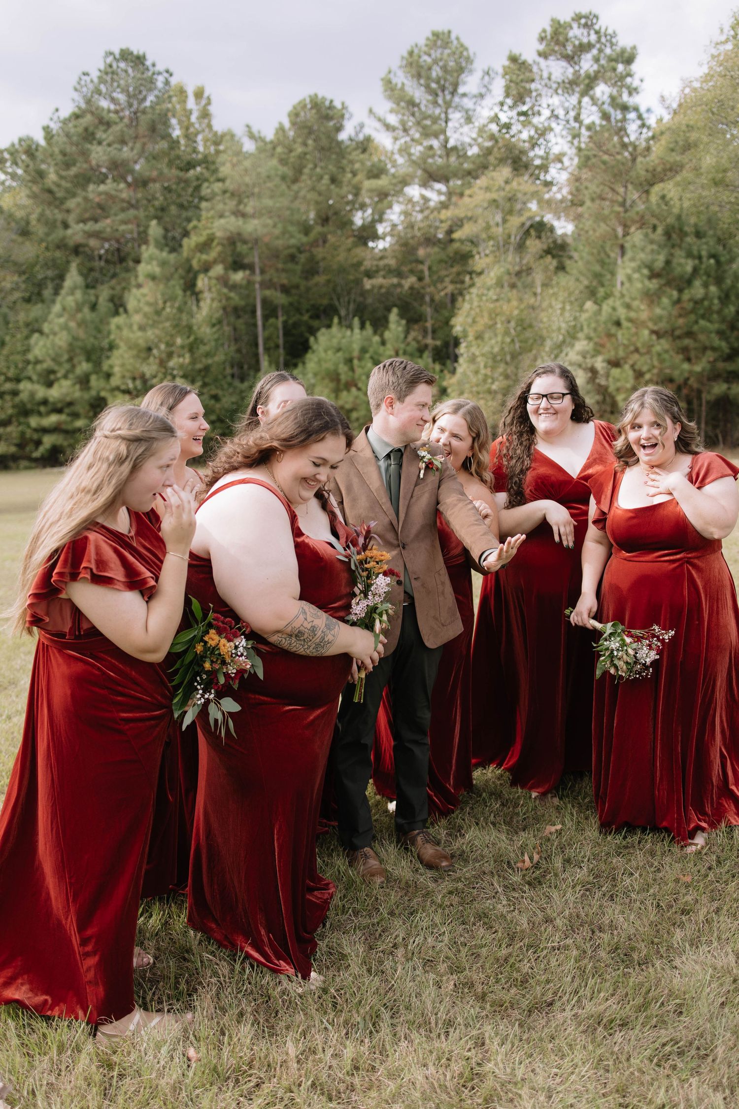 Bridesmaids in red velvet dresses pose together in grassy field.