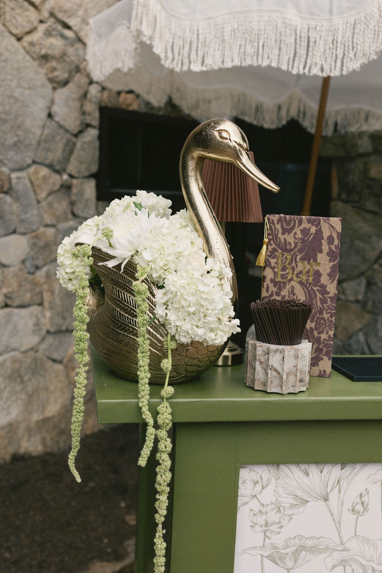 Gold swan vase with white hydrangeas on green table, next to decorative cards and matches, stone wall backdrop.