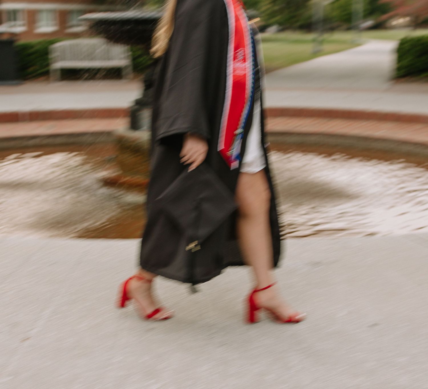 A graduate in black cap and gown with red sash walks in heels on campus near a fountain.