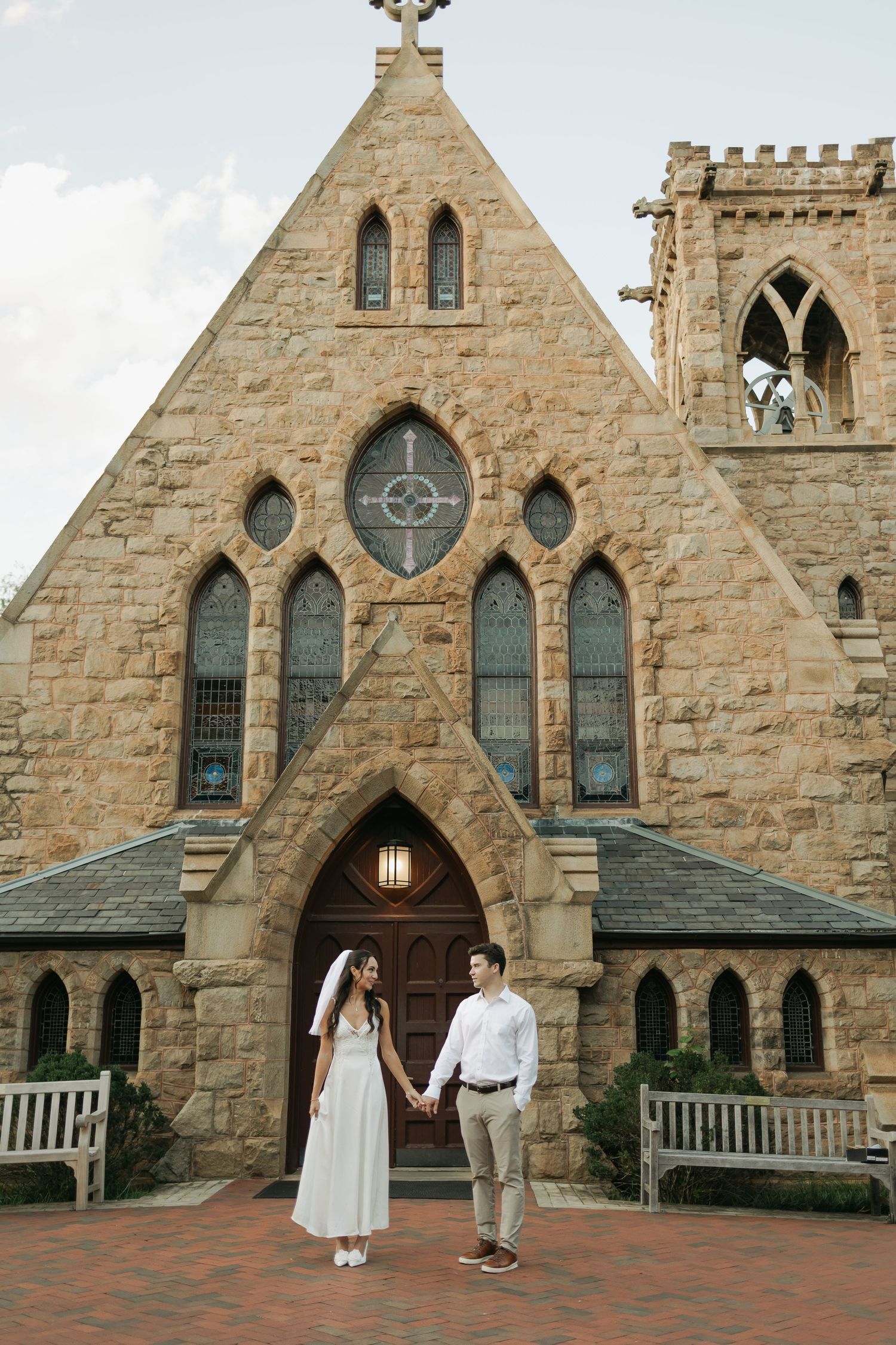 Bride and groom holding hands in front of a historic Gothic stone church with arched windows and brick courtyard.