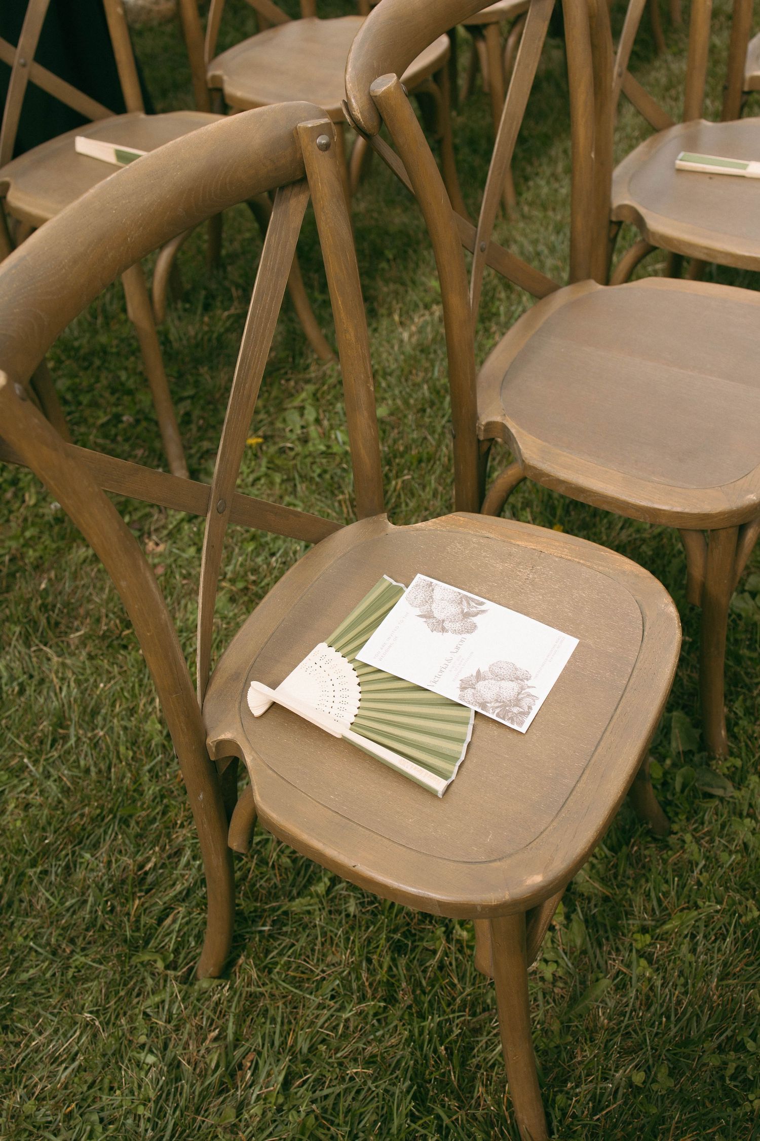 Wooden cross-back chairs on grass with green folding fans and wedding programs placed on seats for outdoor ceremony.