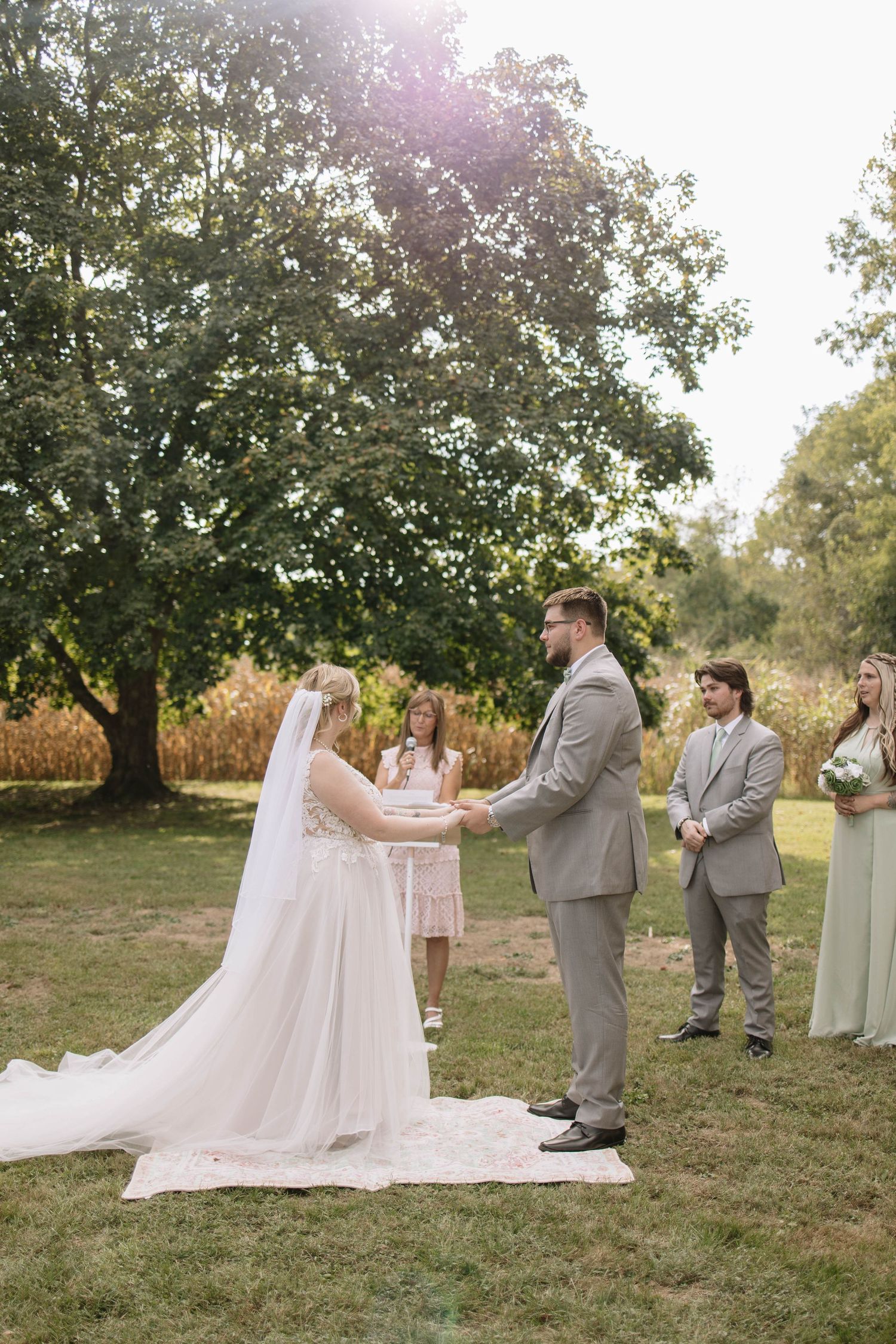 A wedding ceremony taking place on a grassy hillside with attendants in grey and sage green attire.