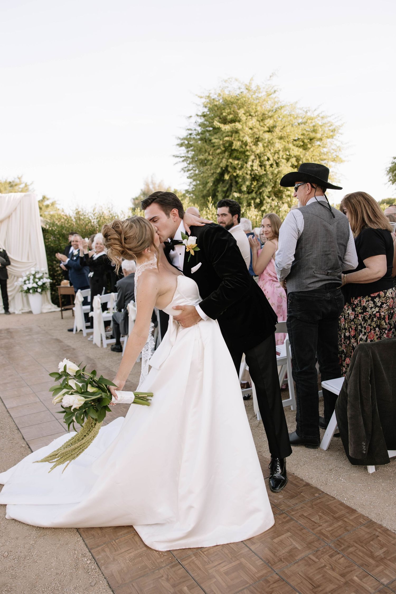 Bride and groom share a kiss at outdoor wedding ceremony while guests look on, bride holding white floral bouquet.