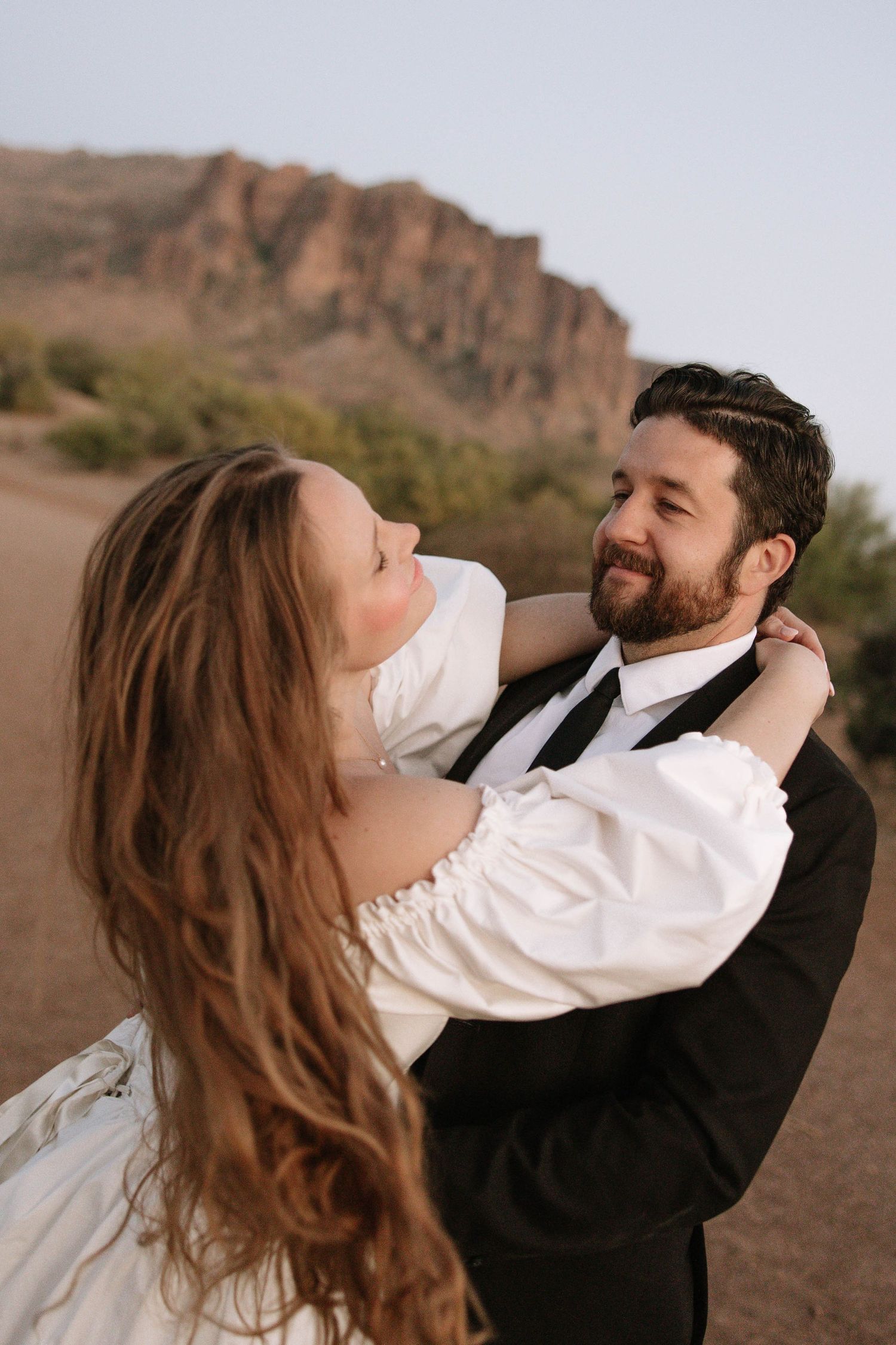 A bride and groom share a romantic embrace outdoors with a stunning red rock mountain backdrop on their wedding day.