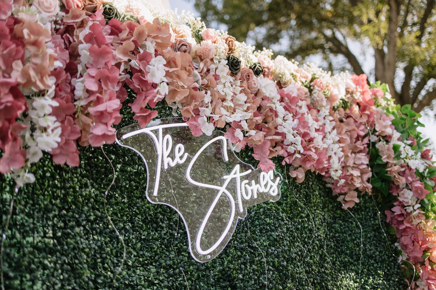 An elegant wedding ceremony setup with pink floral arrangements and a neon sign against a green hedge wall.