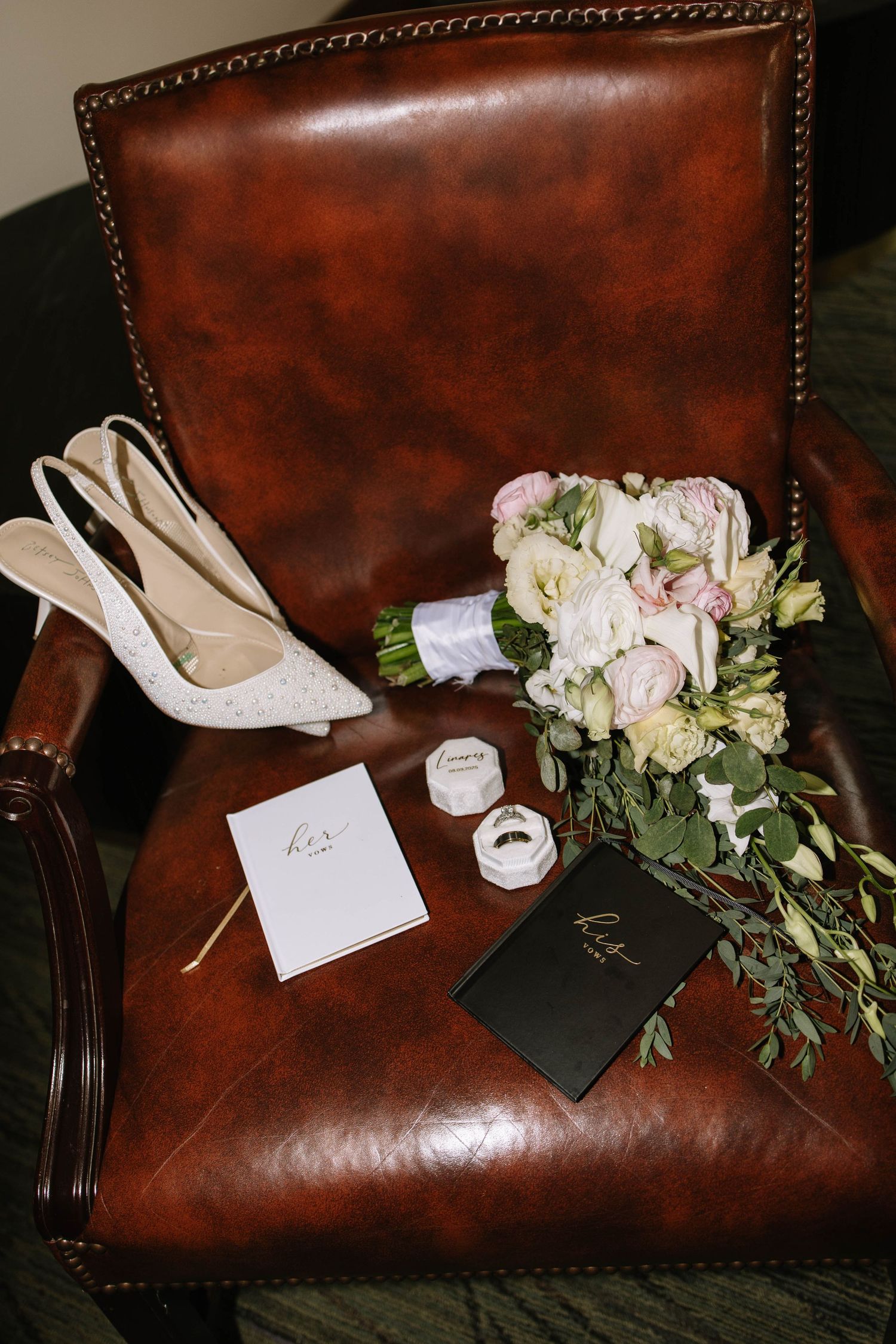 Wedding accessories including white heels and pink flowers arranged on brown leather couch.