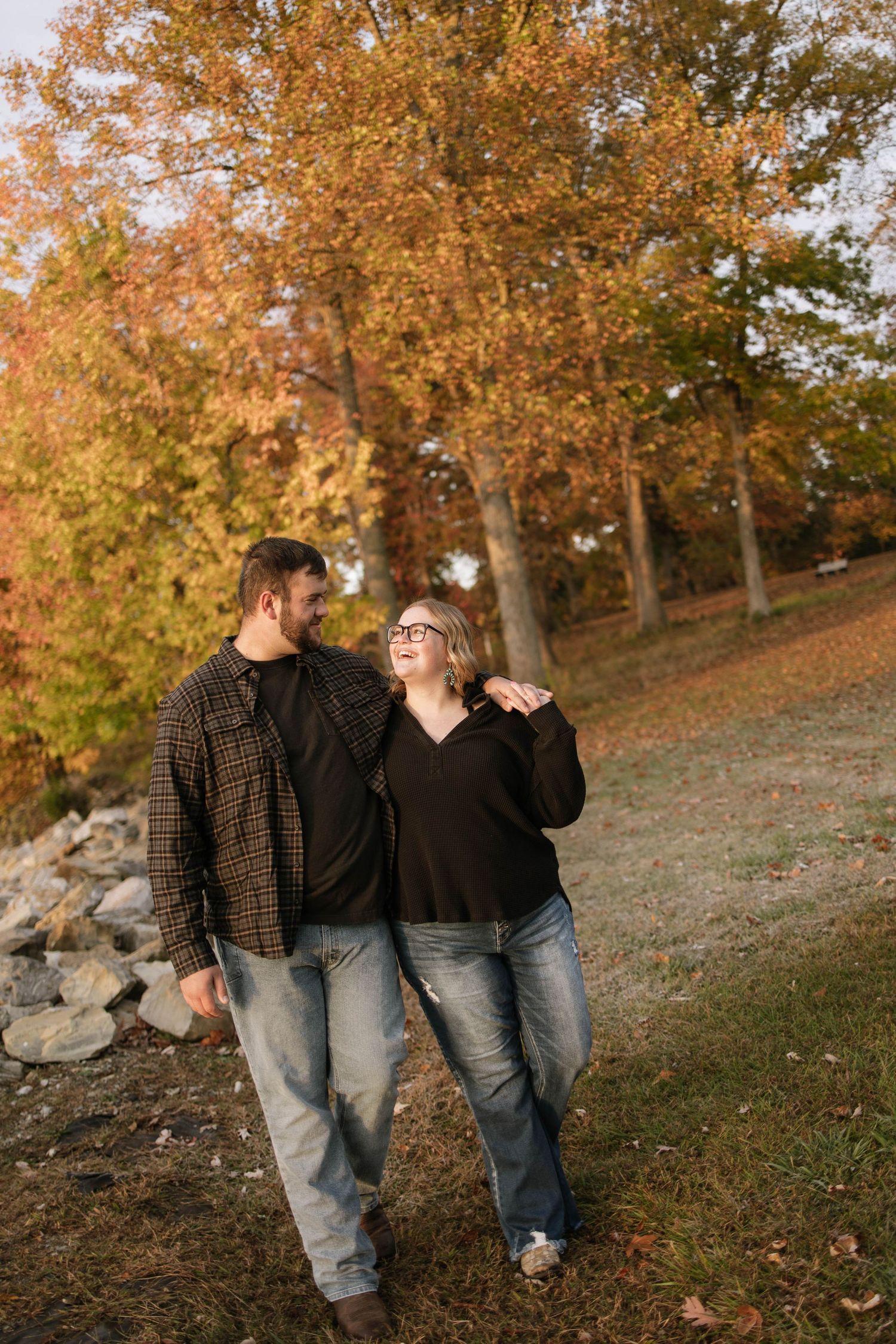 A couple walks together through a park with autumn trees and fallen leaves in the background.