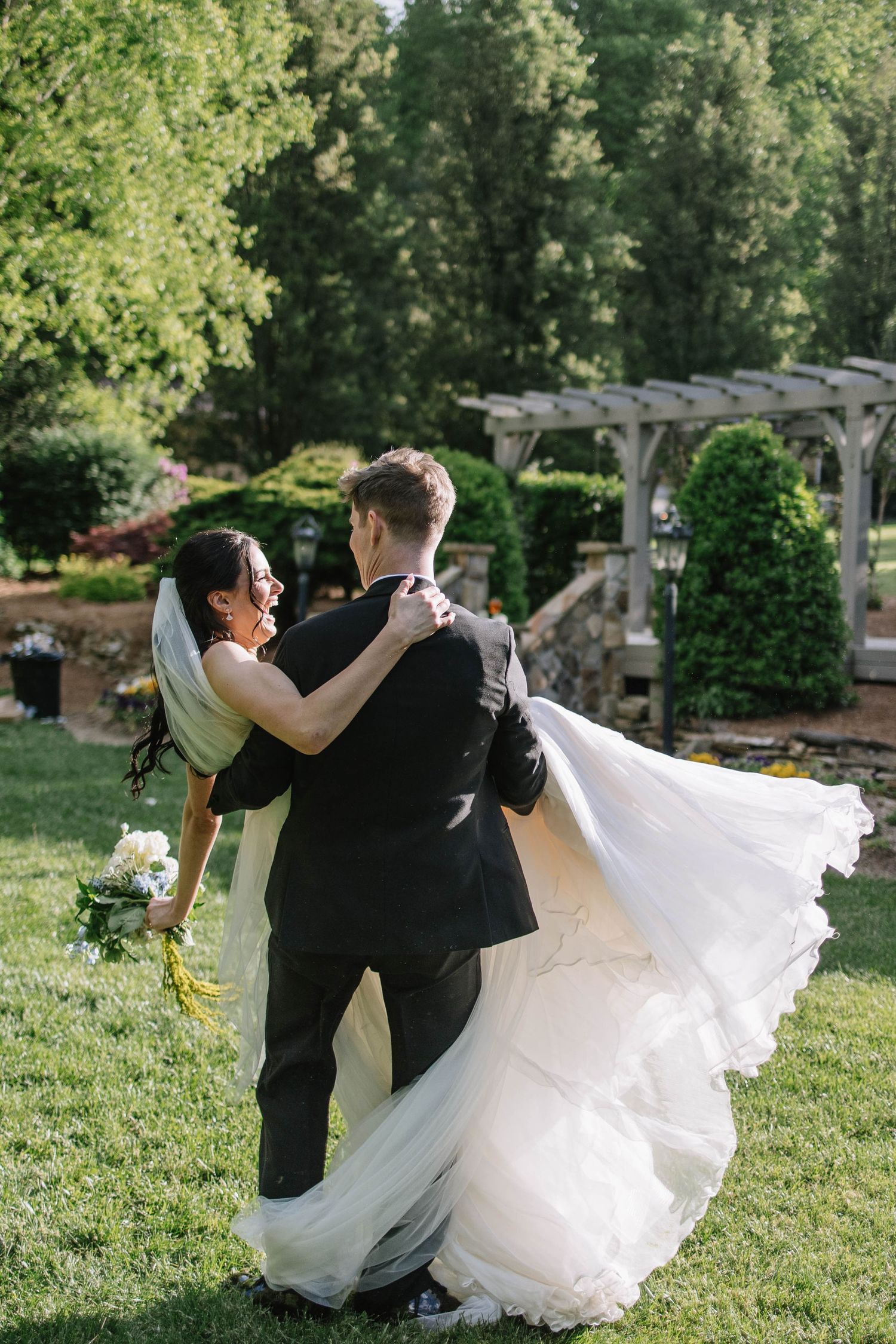A joyful bride in a flowing white gown and groom in a black suit dance outdoors in a lush green garden setting.