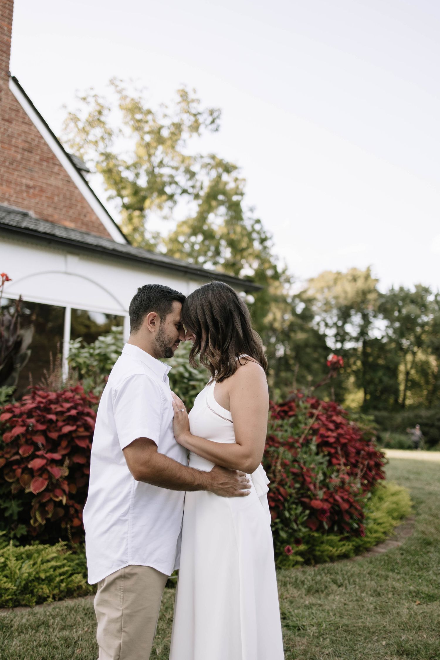 A couple in coordinating white outfits embraces near red foliage and a brick building.