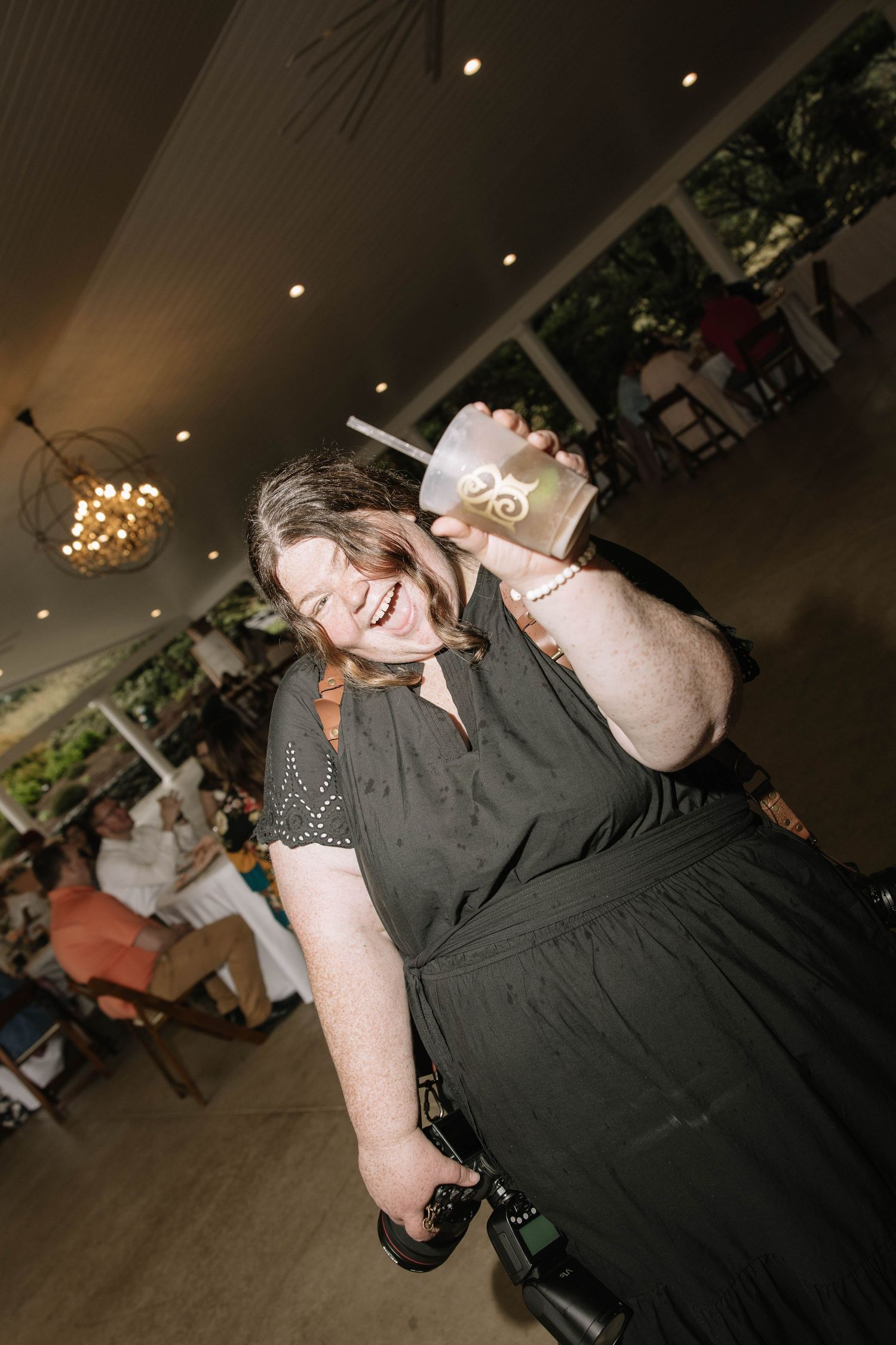 Woman in black dress laughing joyfully while holding a drink at an indoor event venue.