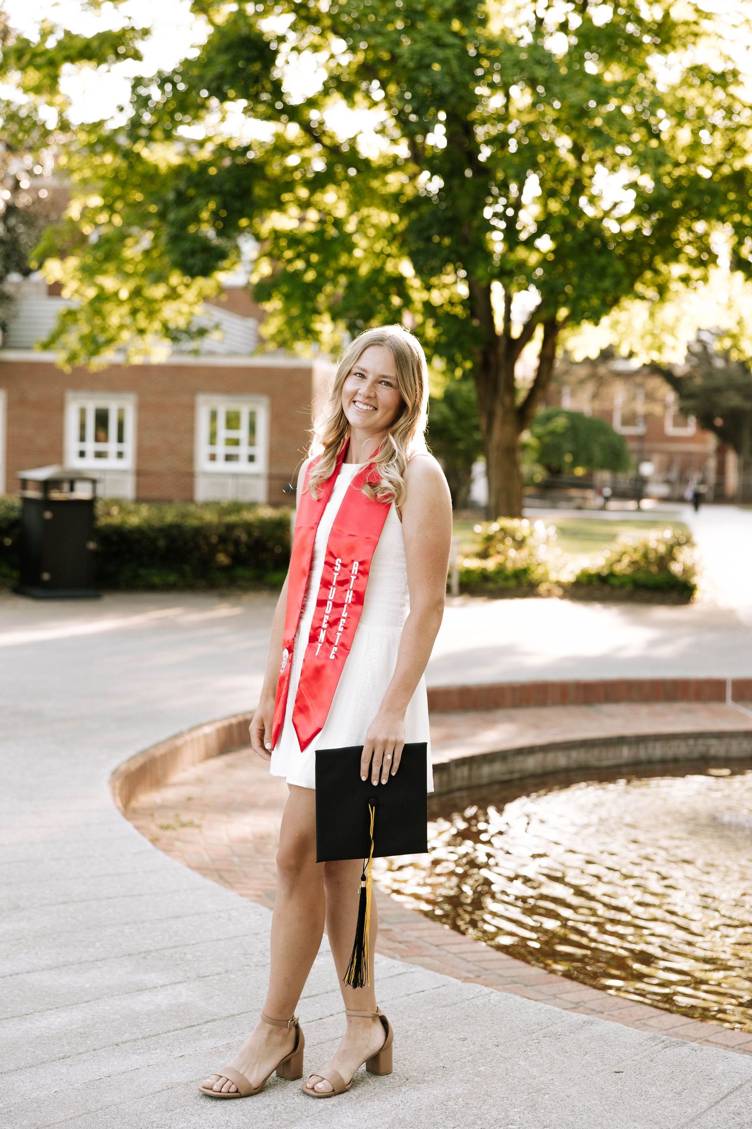Smiling female graduate in white dress with red sash holds cap near fountain on sunny campus.
