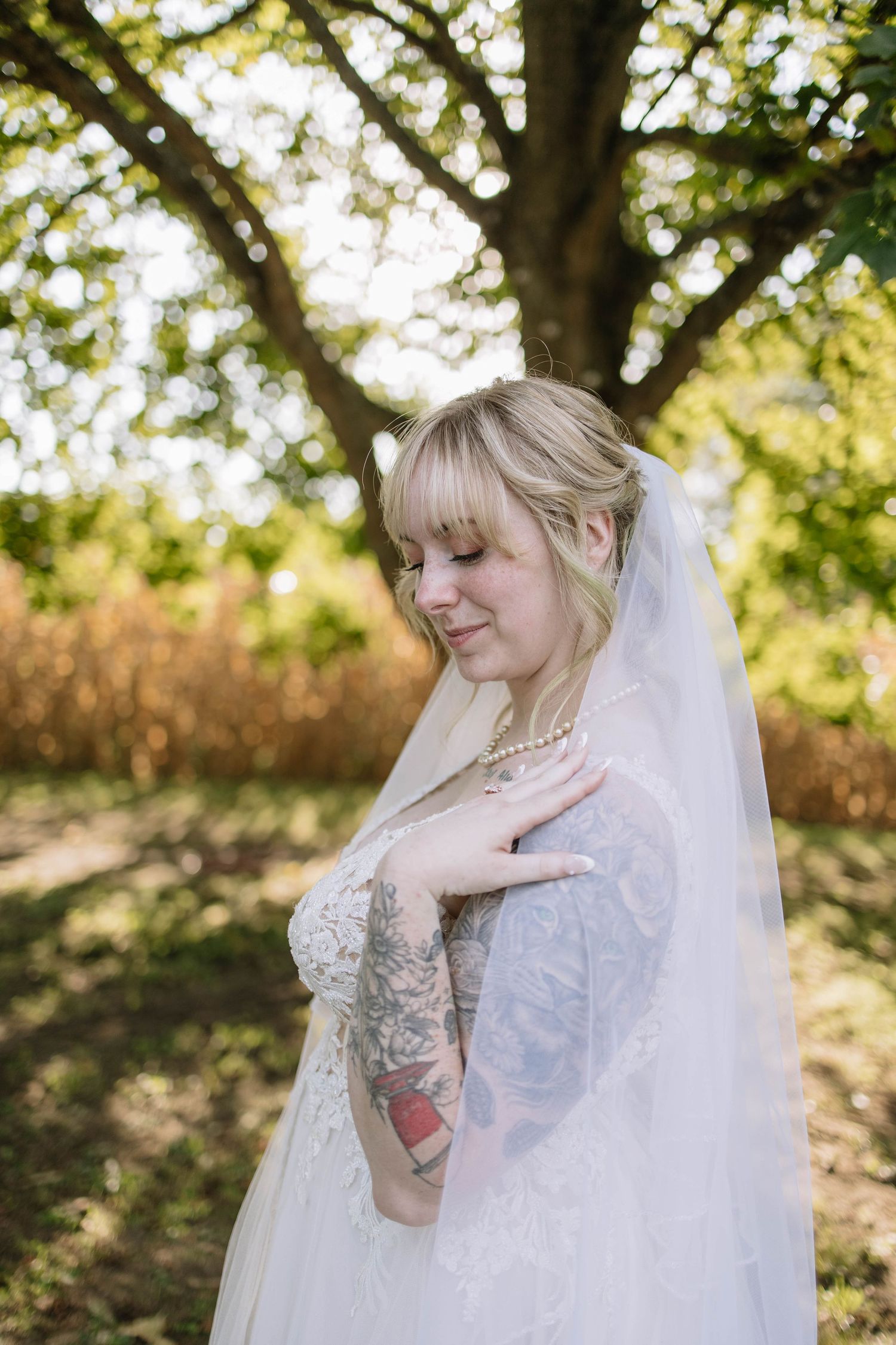 Bride in lace dress with flowing veil poses outdoors against natural foliage.