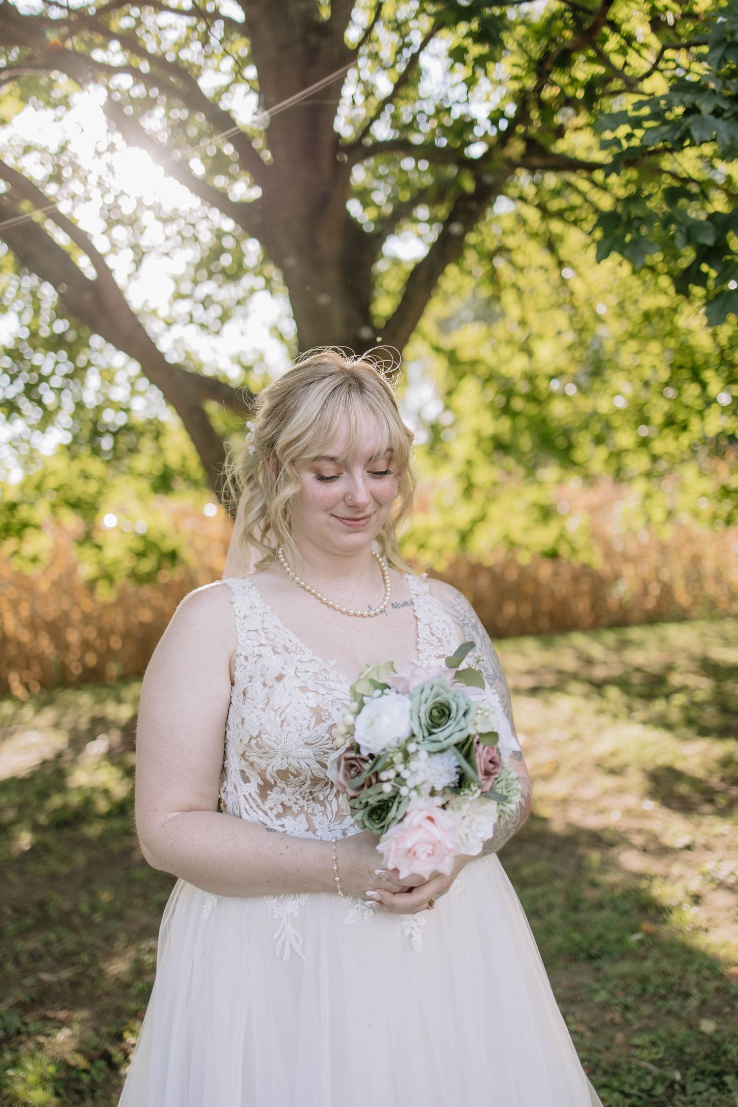 Bride in white lace gown holding delicate bouquet poses in natural outdoor setting.