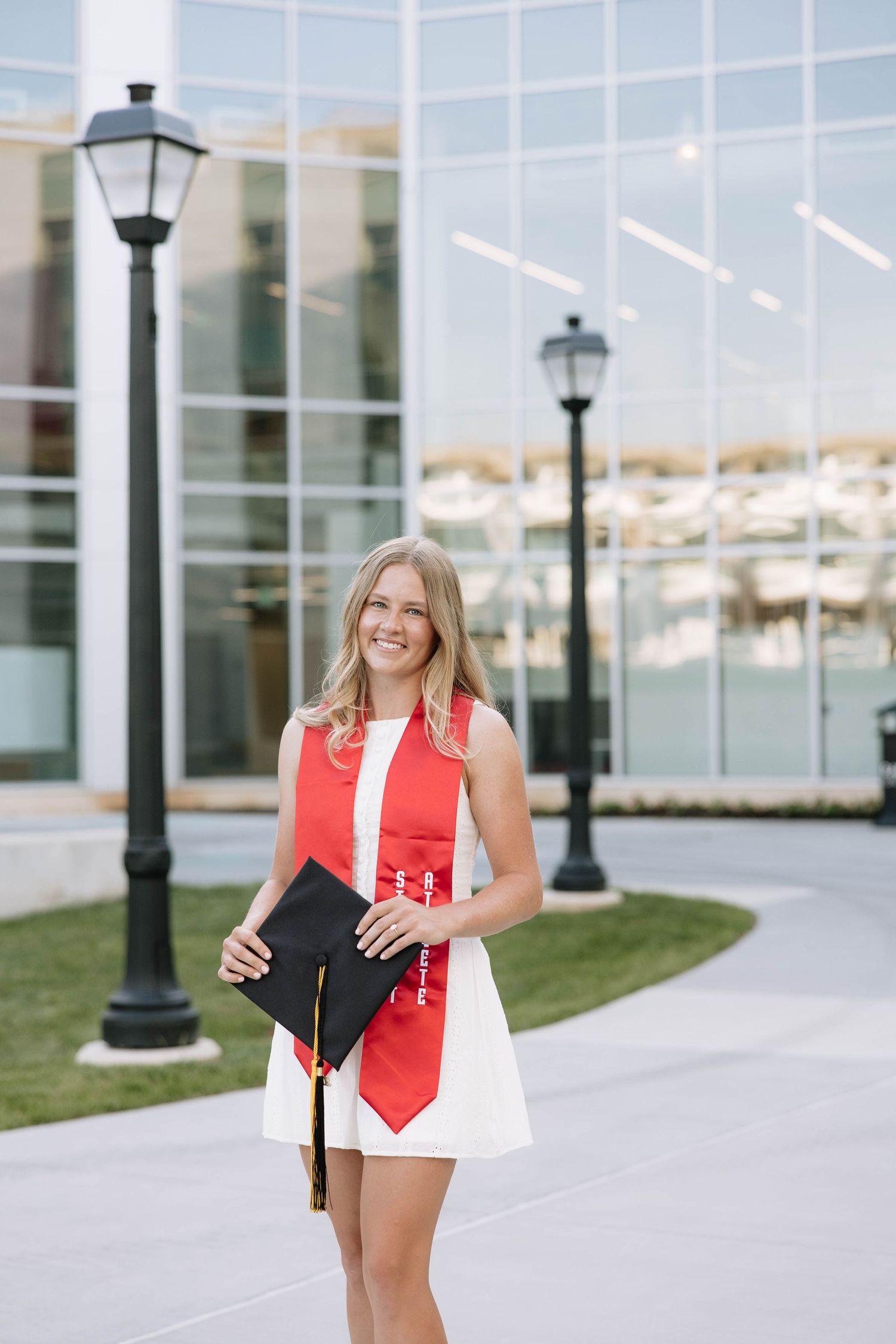 Female graduate in white dress and red honor stole holds cap while smiling outside modern university building.