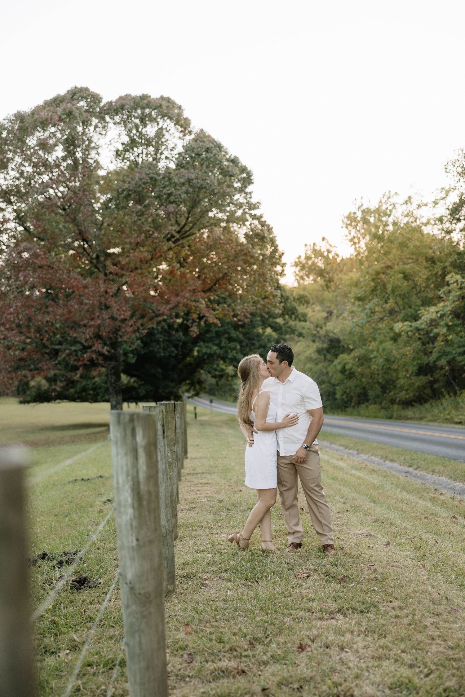 A couple shares an intimate moment on a grassy hill with trees and a winding path in the background.
