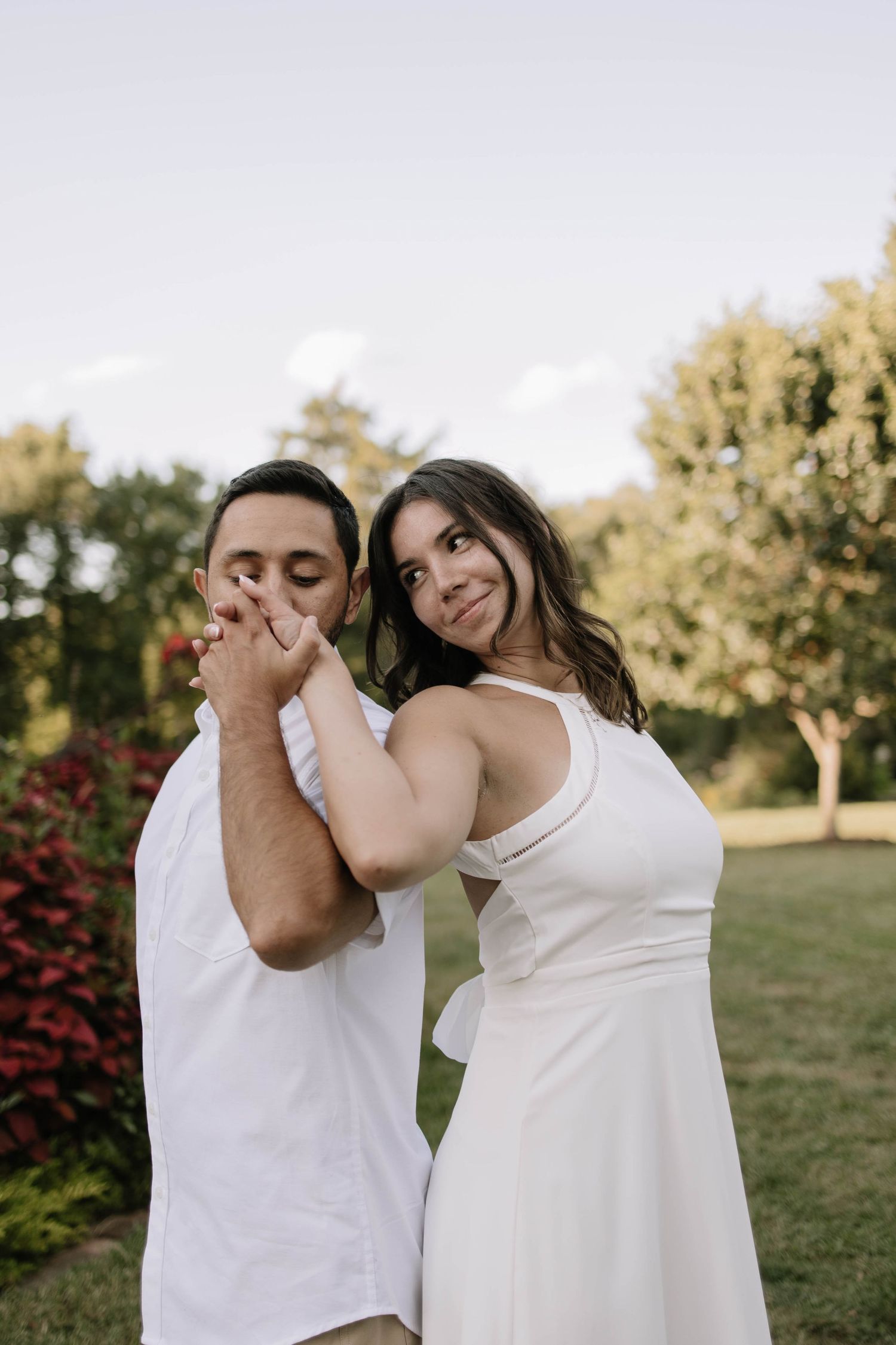 A couple in white outfits dances together on a grassy lawn with red bushes in the background.