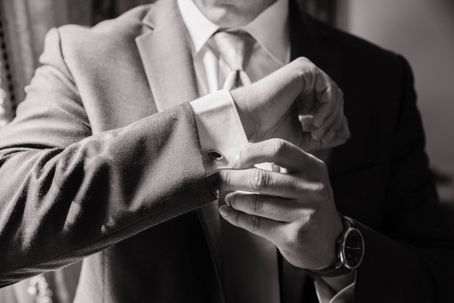 Close-up black and white image of someone adjusting their shirt cuff while wearing a formal gray suit and wristwatch.