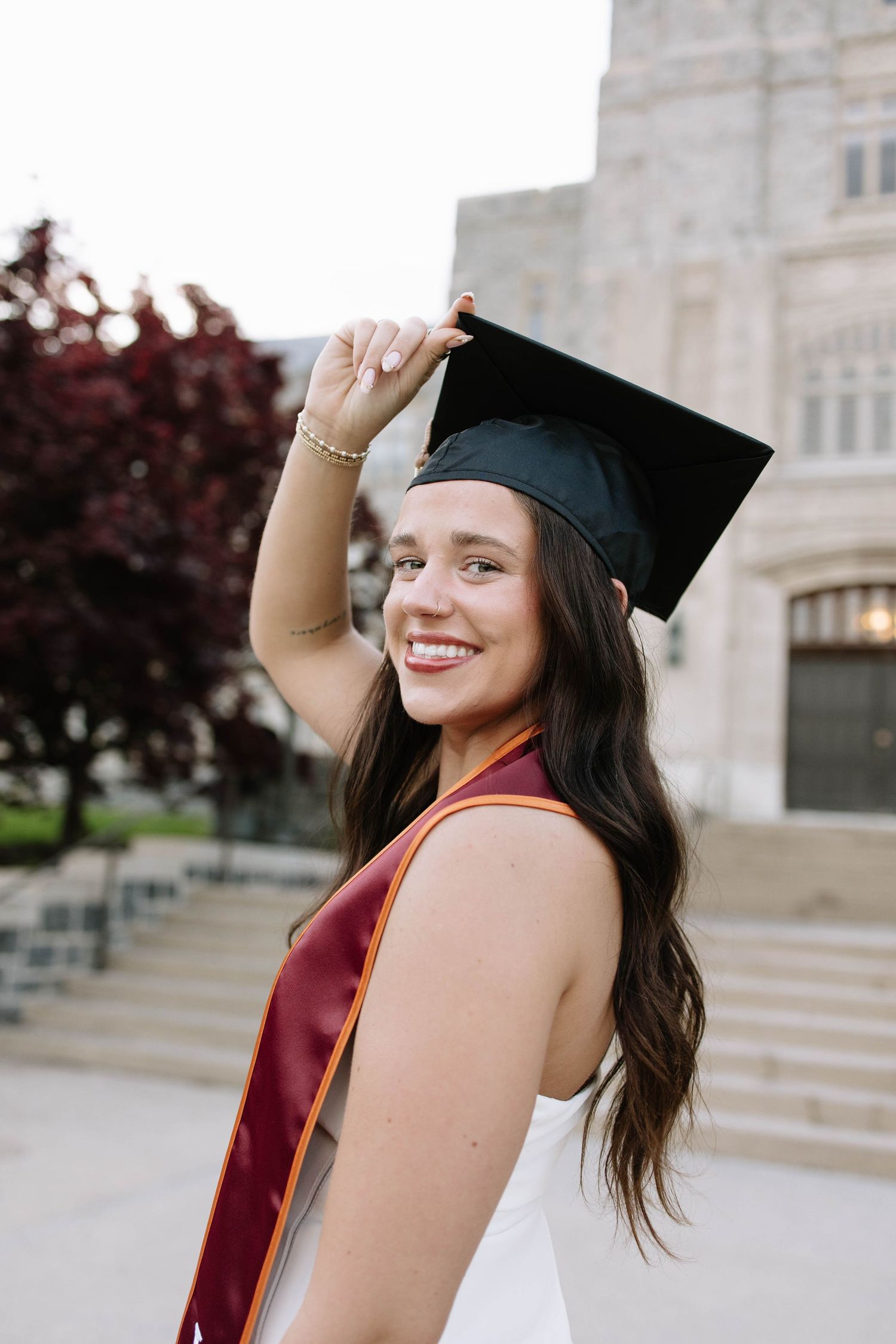 Smiling young woman in black graduation cap and red sash poses outdoors near university building steps.