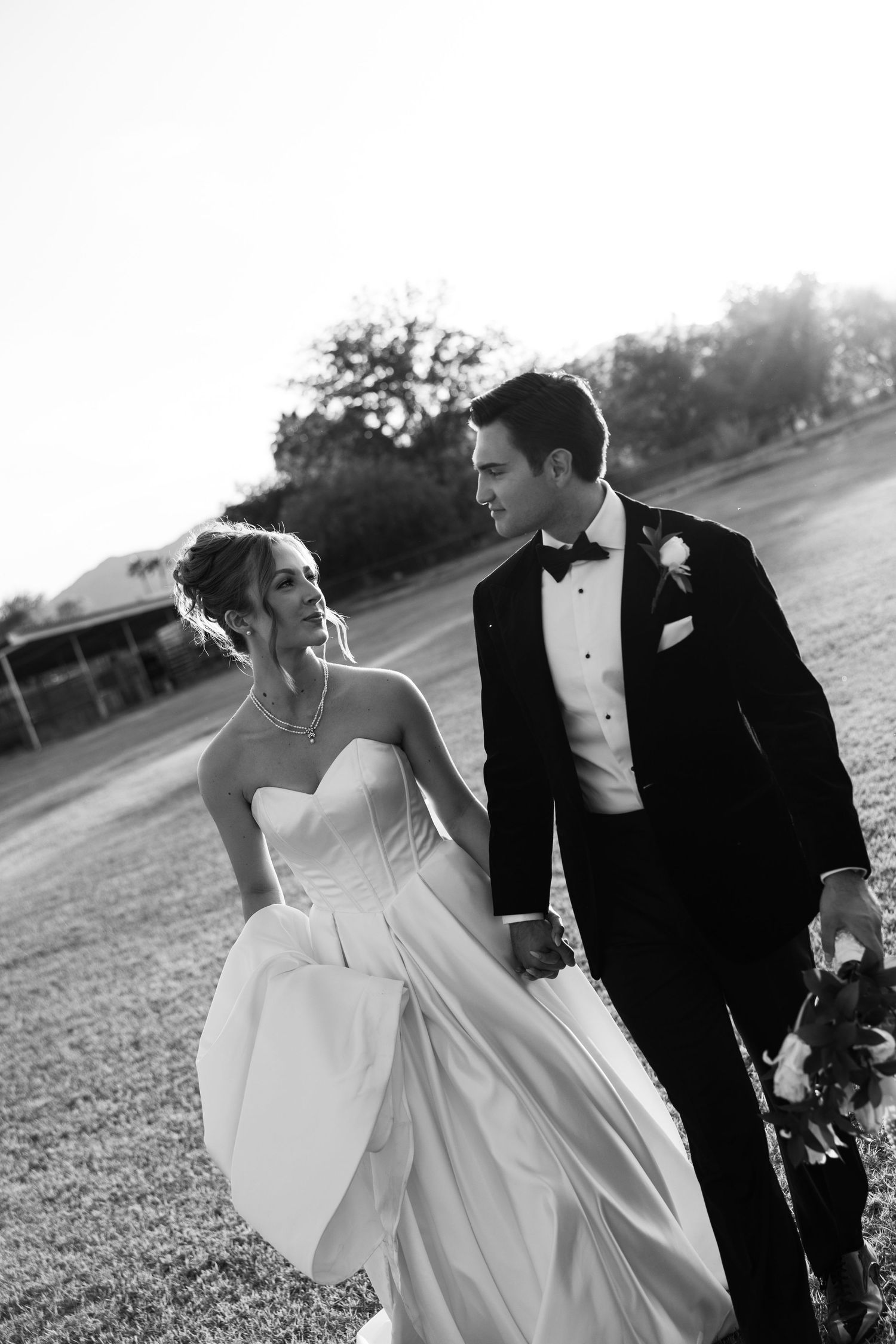 Black and white photo of a bride and groom walking hand in hand outdoors, gazing at each other on their wedding day.