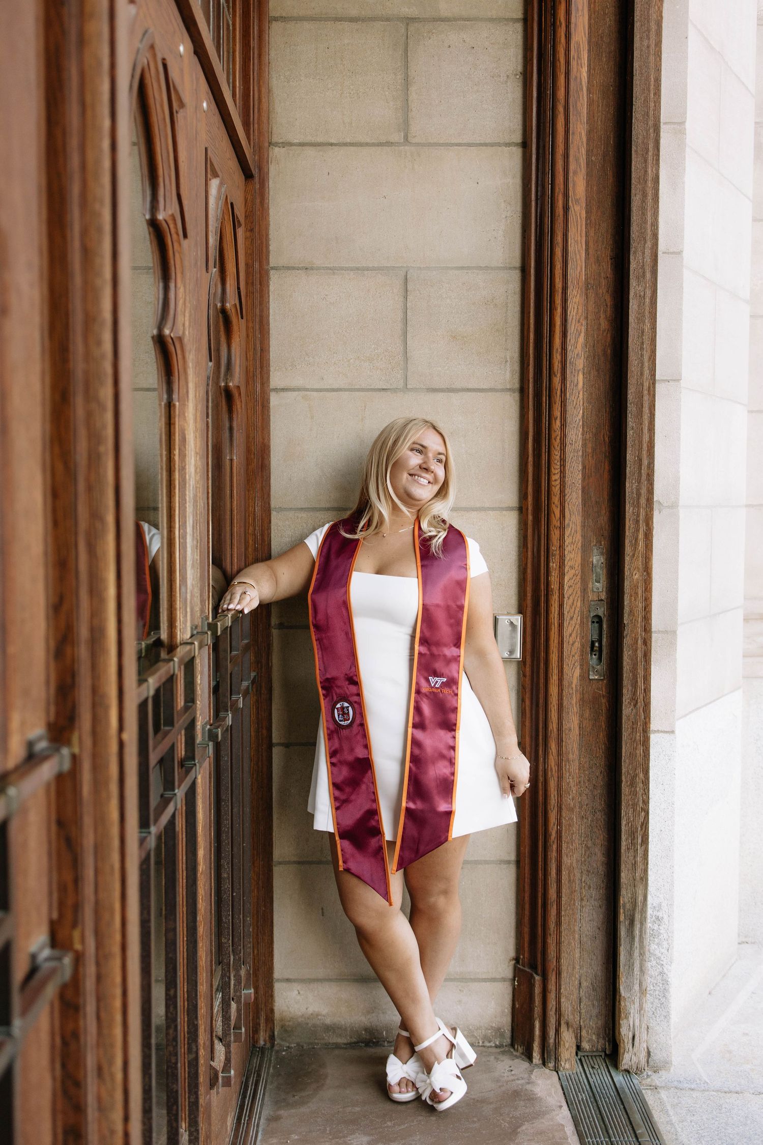 Smiling graduate in white dress and maroon stole poses between wooden doors at graduation photo session.