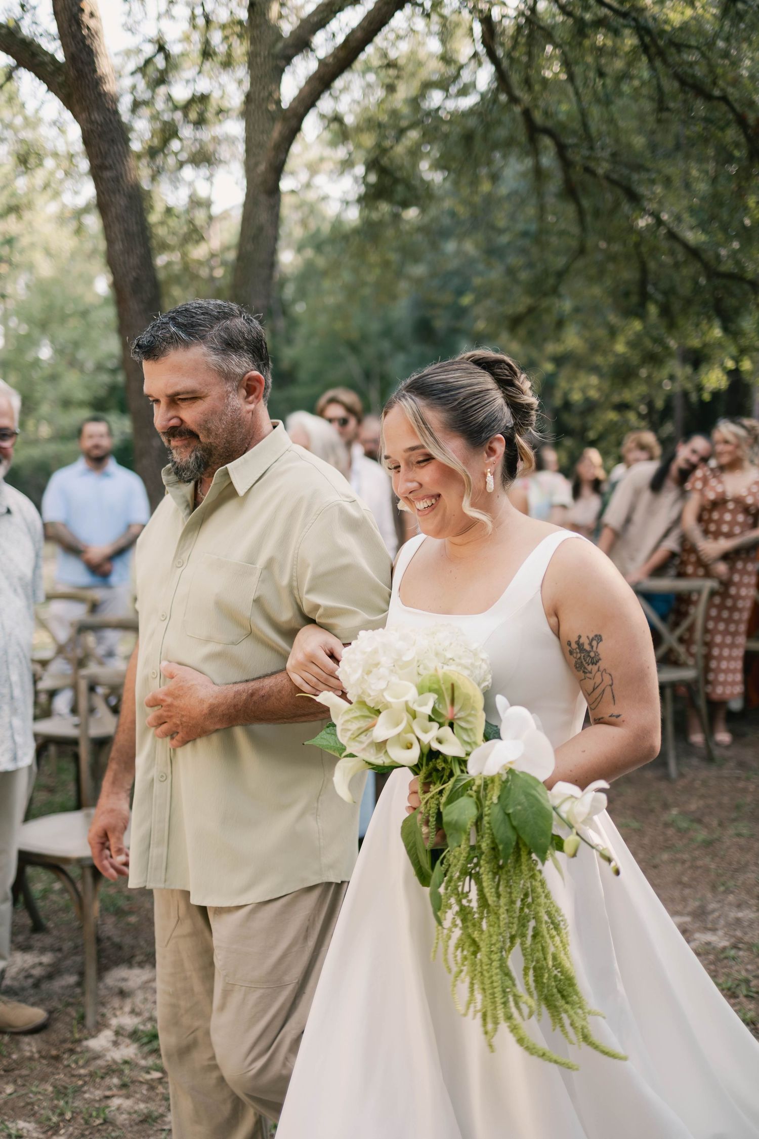 A smiling bride in a white gown holds a white floral bouquet while walking down the aisle with a bearded man.