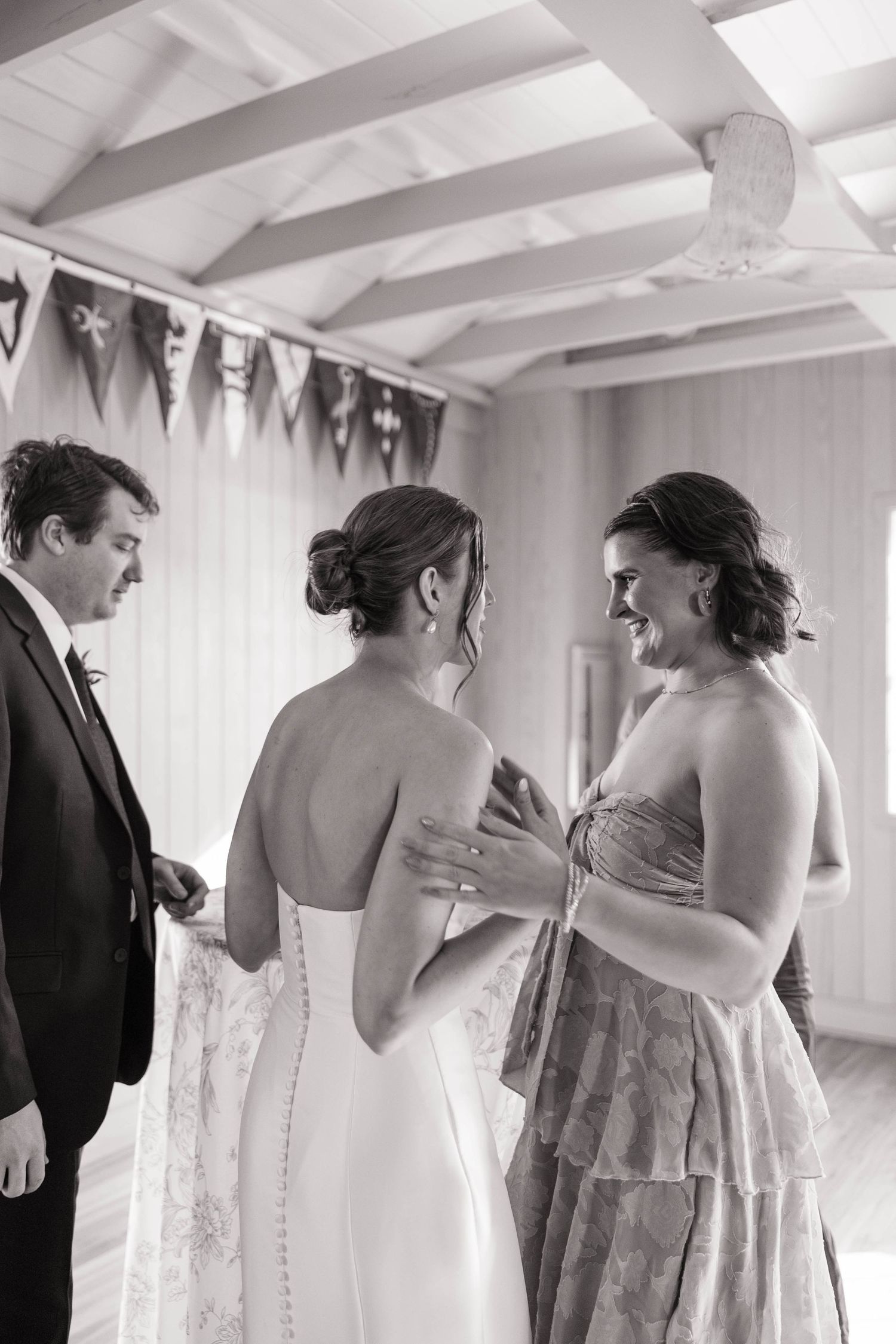 Black and white photo of a bride in a backless gown being greeted by a smiling woman at a wedding reception.