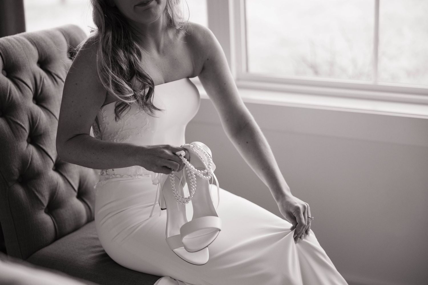 A bride in a white dress sits elegantly on a tufted chair near a window in black and white photography.